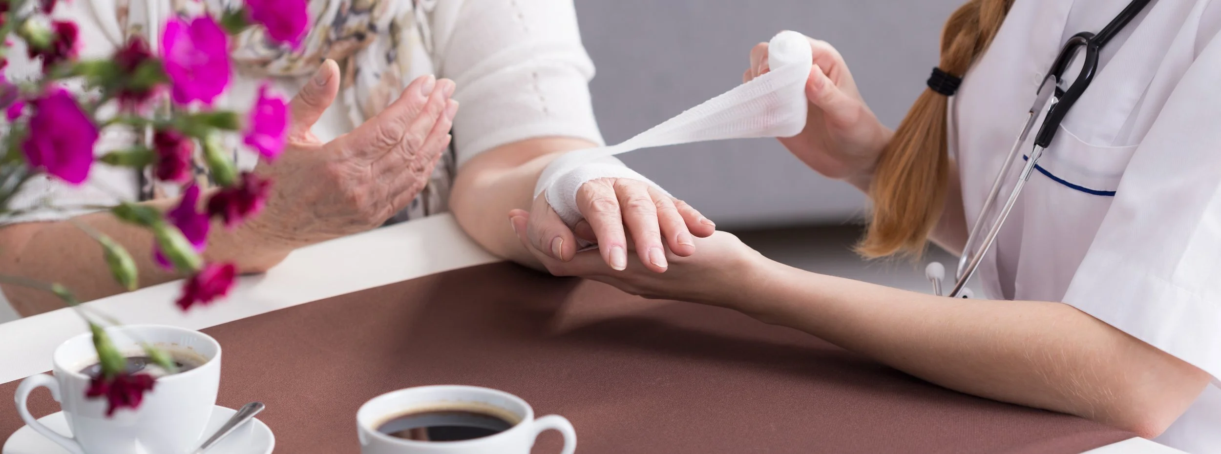 A healthcare worker assesses an elderly patient's arm, which is wrapped in bandages, in a clinical setting. There are two cups of coffee and a vase with pink flowers on the table.