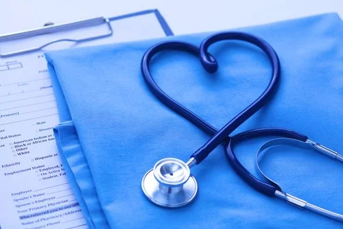 A stethoscope with its tubing shaped into a heart on blue medical scrubs, placed next to a clipboard with medical documents.