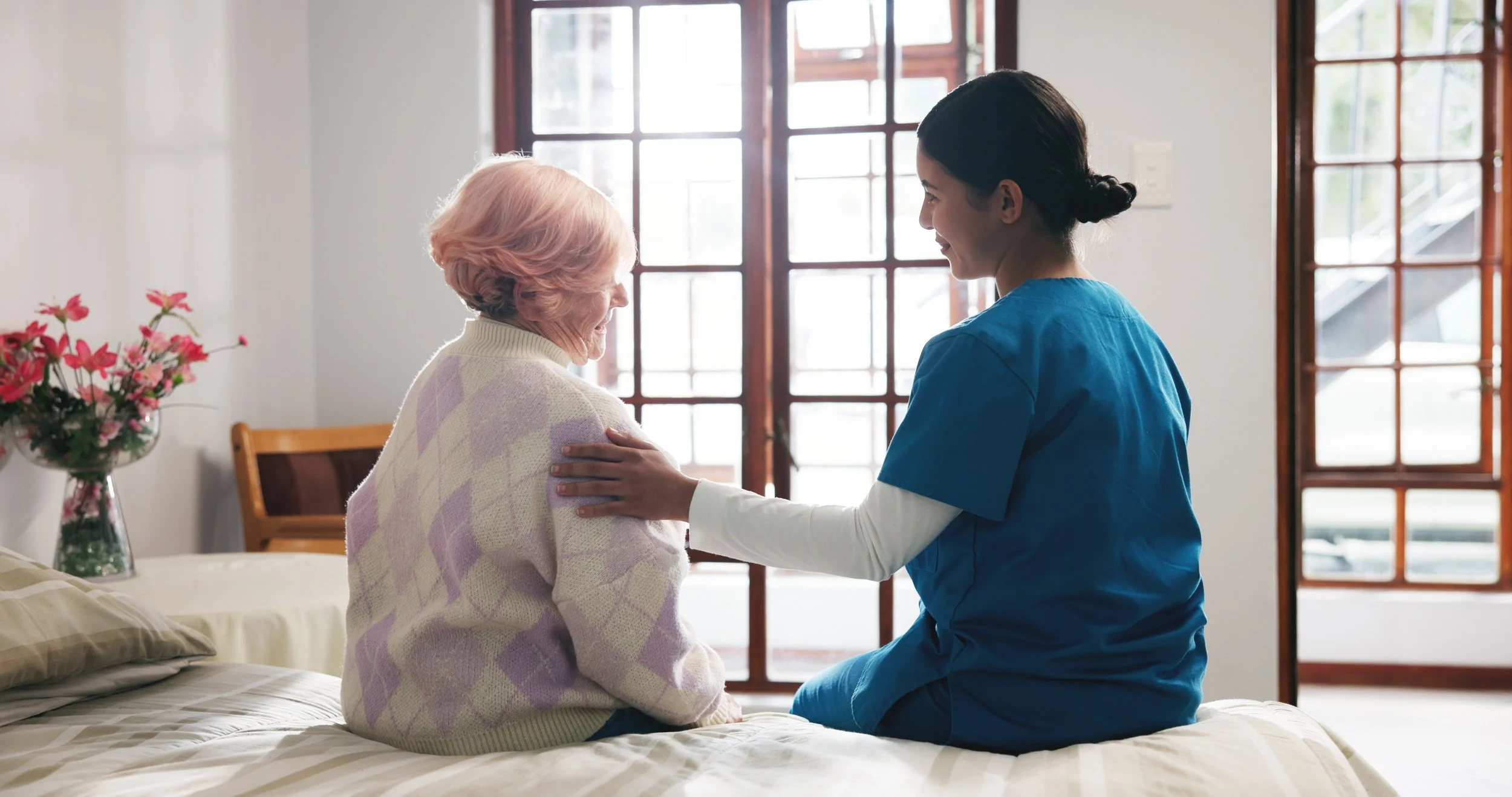 A caregiver in scrubs showing compassion to an elderly woman sitting on a bed in a well-lit room.