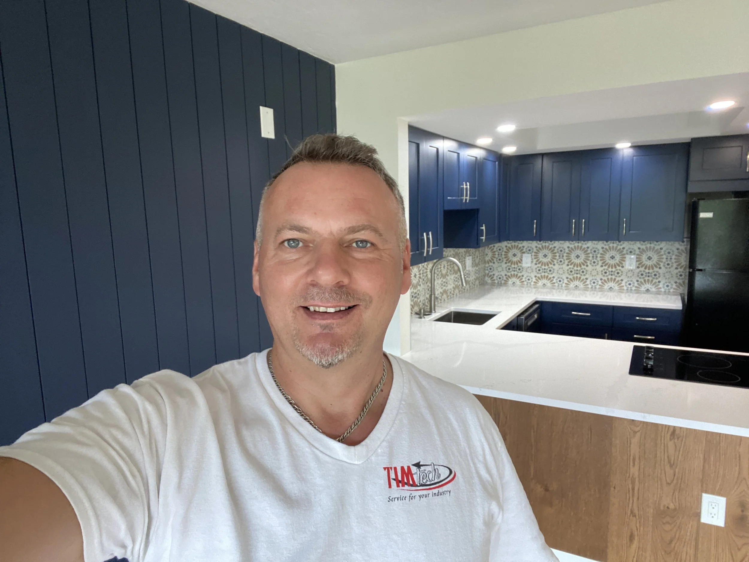 A smiling man taking a selfie in a modern kitchen with blue cabinets, a white countertop, and a patterned tile backsplash.