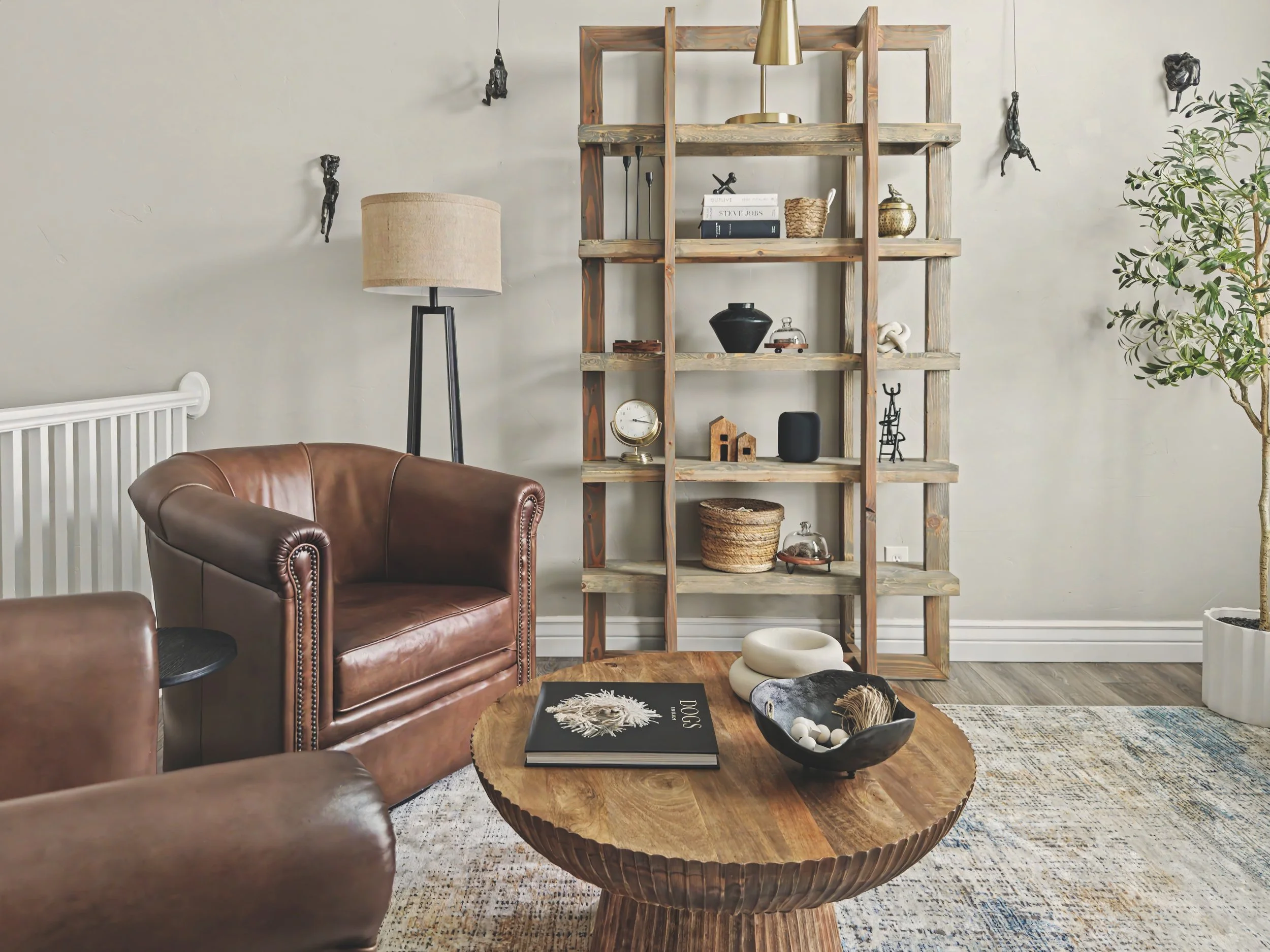 A cozy living room with a brown leather armchair and a wooden coffee table holding a large book titled "Dogs" and a black bowl filled with decorative objects. In the background, a tall wooden bookshelf displays books, vases, and decorative items, next to a potted plant with green and white leaves. The wall behind features black hanging sculptures, and a beige floor lamp with a black tripod base is beside the bookshelf. There is a textured rug under the coffee table and a striped rug partially visible on the floor.
