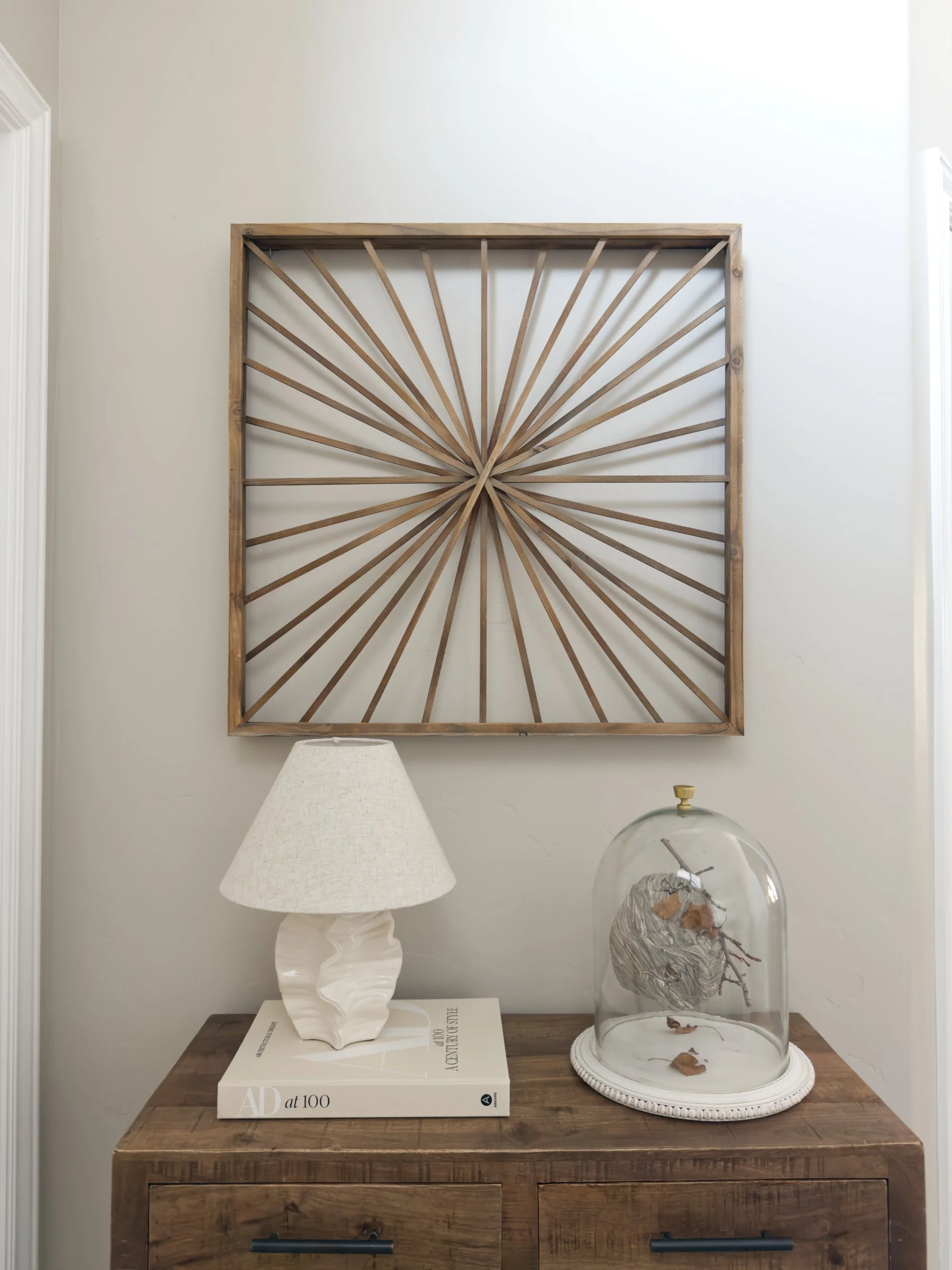 Decorative wooden wall art with a sunburst pattern mounted above a wooden sideboard. The sideboard holds a white textured table lamp on a book and a glass dome encasing a driftwood piece with leaves inside.