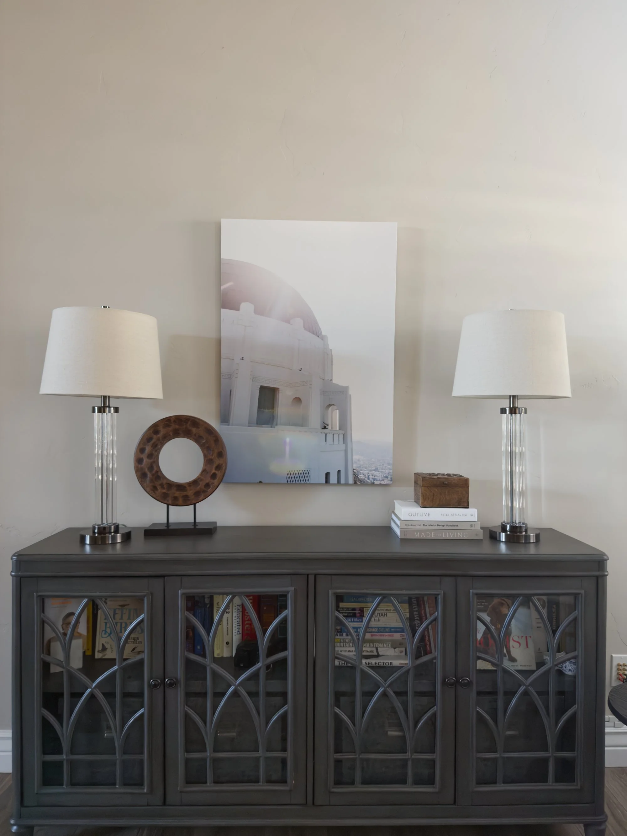 Custom decorated gray sideboard in Bountiful, UT, with glass-paneled doors, topped with two white table lamps, a wooden art piece, a stack of books, and a framed photo of a white building with a dome.