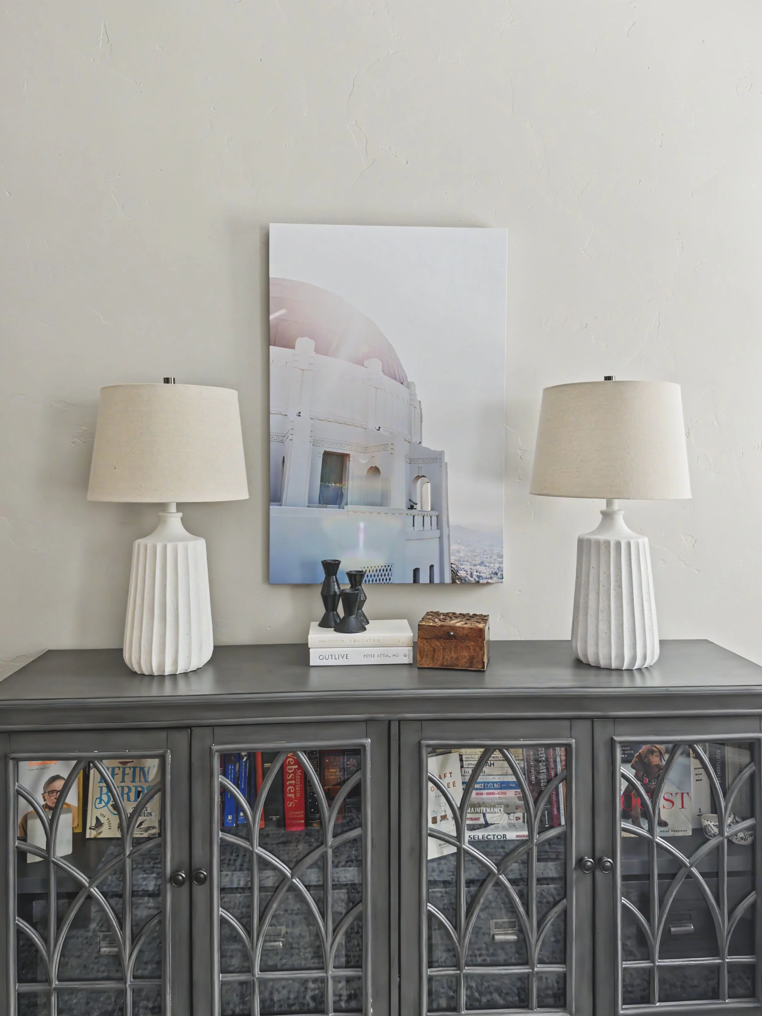 A gray sideboard with lattice glass doors holding books and magazines. On top are two white lamps with pleated shades, a small mannequin, and other decorative objects. A large framed photograph of a white building with a glass dome is mounted on the wall behind the sideboard.