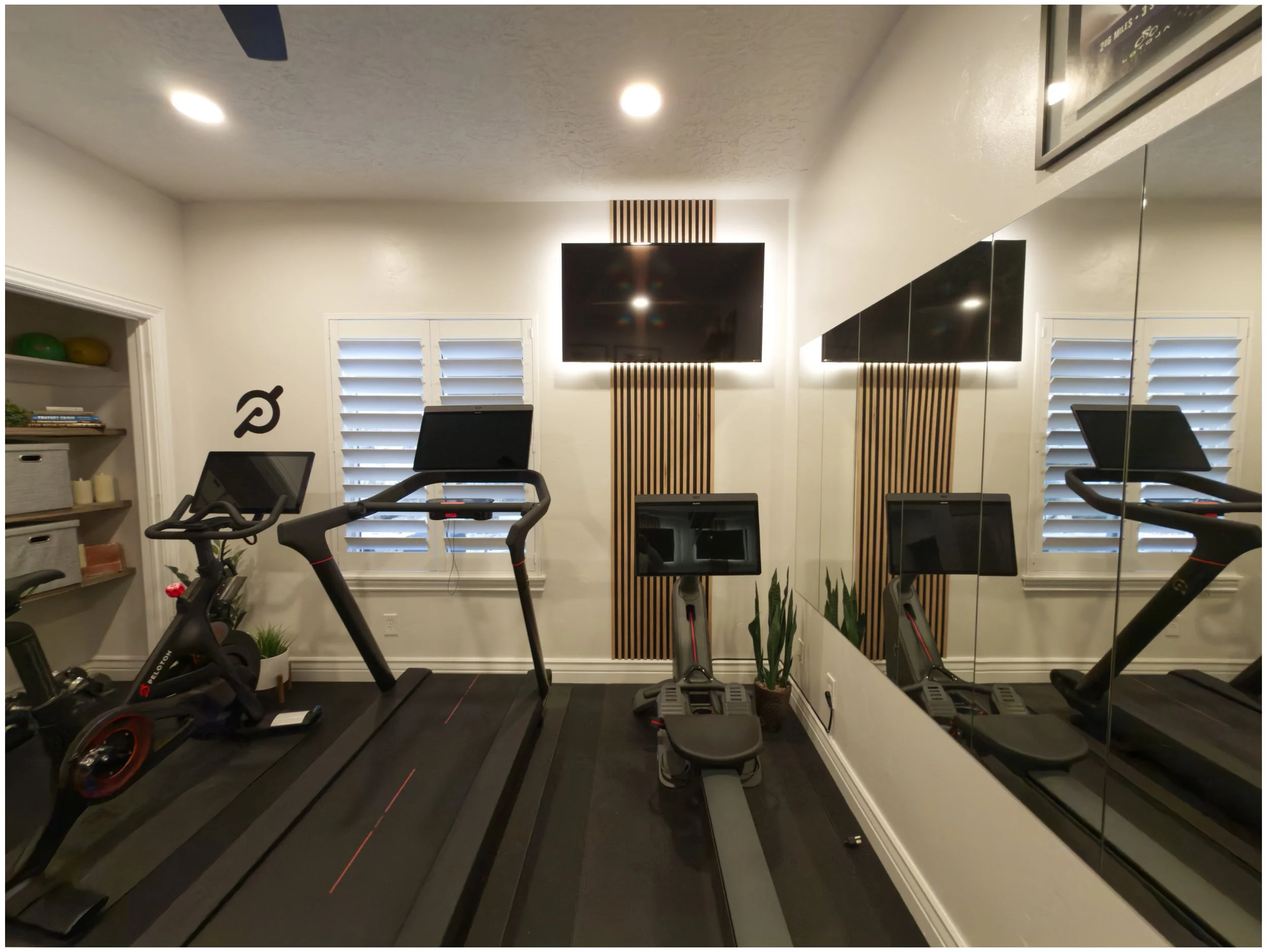 Custom decorated home gym in Salt Lake City, UT with treadmill, stationary bike, and rowing machine in front of a mirror; TV on wall with wooden panel accents; shelves with storage bins and decorative items.