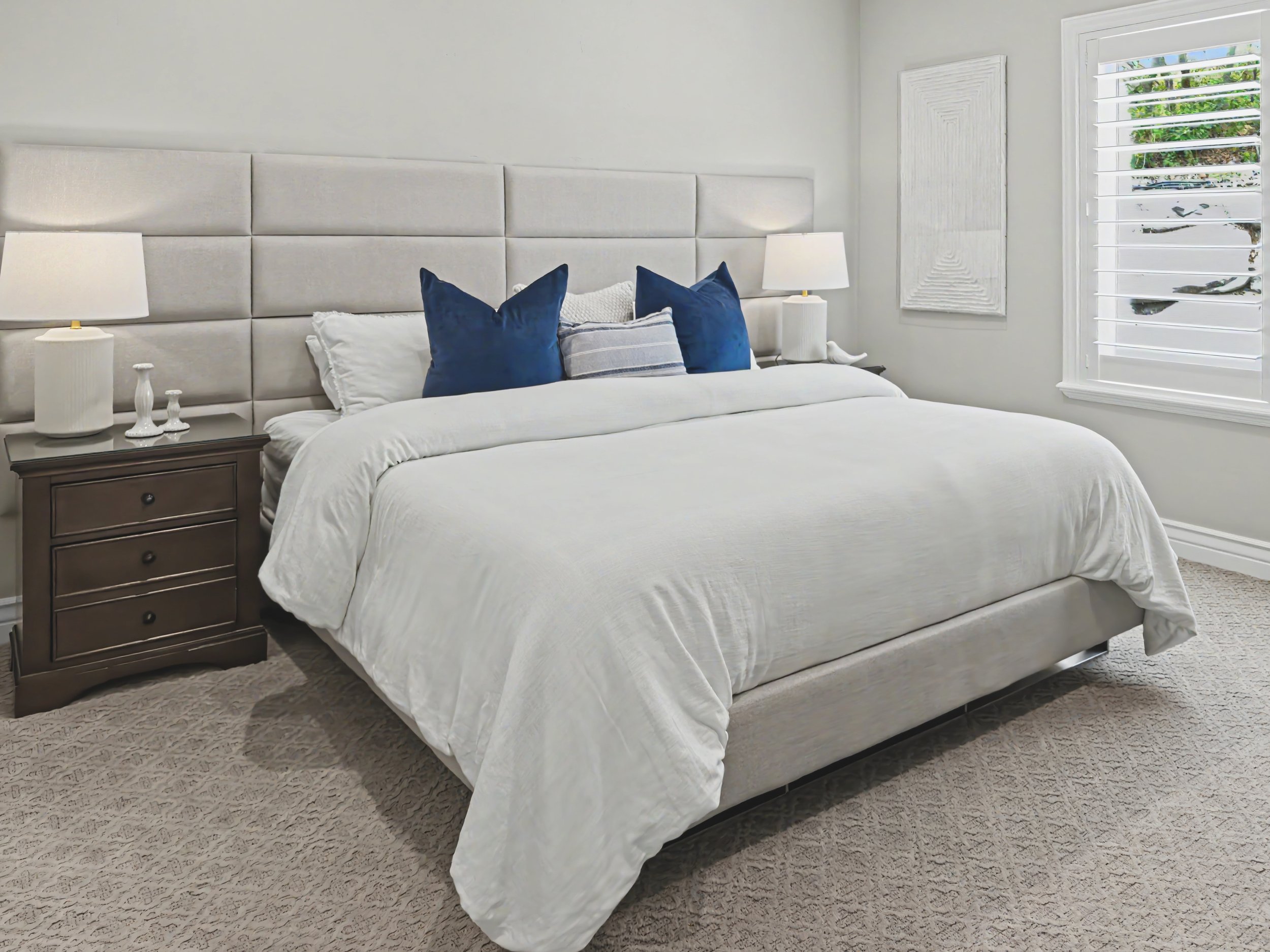 A neatly made bed in a bedroom with a white bedspread and navy blue pillows. There are two white nightstands on either side of the bed, each with a white lamp. The room has a window with white shutters and a view of greenery outside. The walls are painted light gray, and there is a textured white wall art piece next to the window. The floor is covered with a beige carpet.