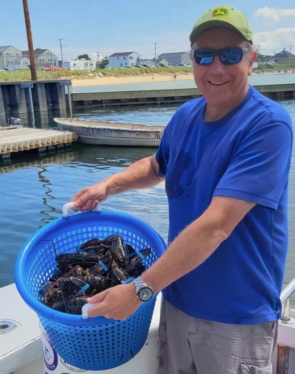 A man in a blue shirt, sunglasses, and a green cap is holding a blue basket filled with lobsters near a dock.