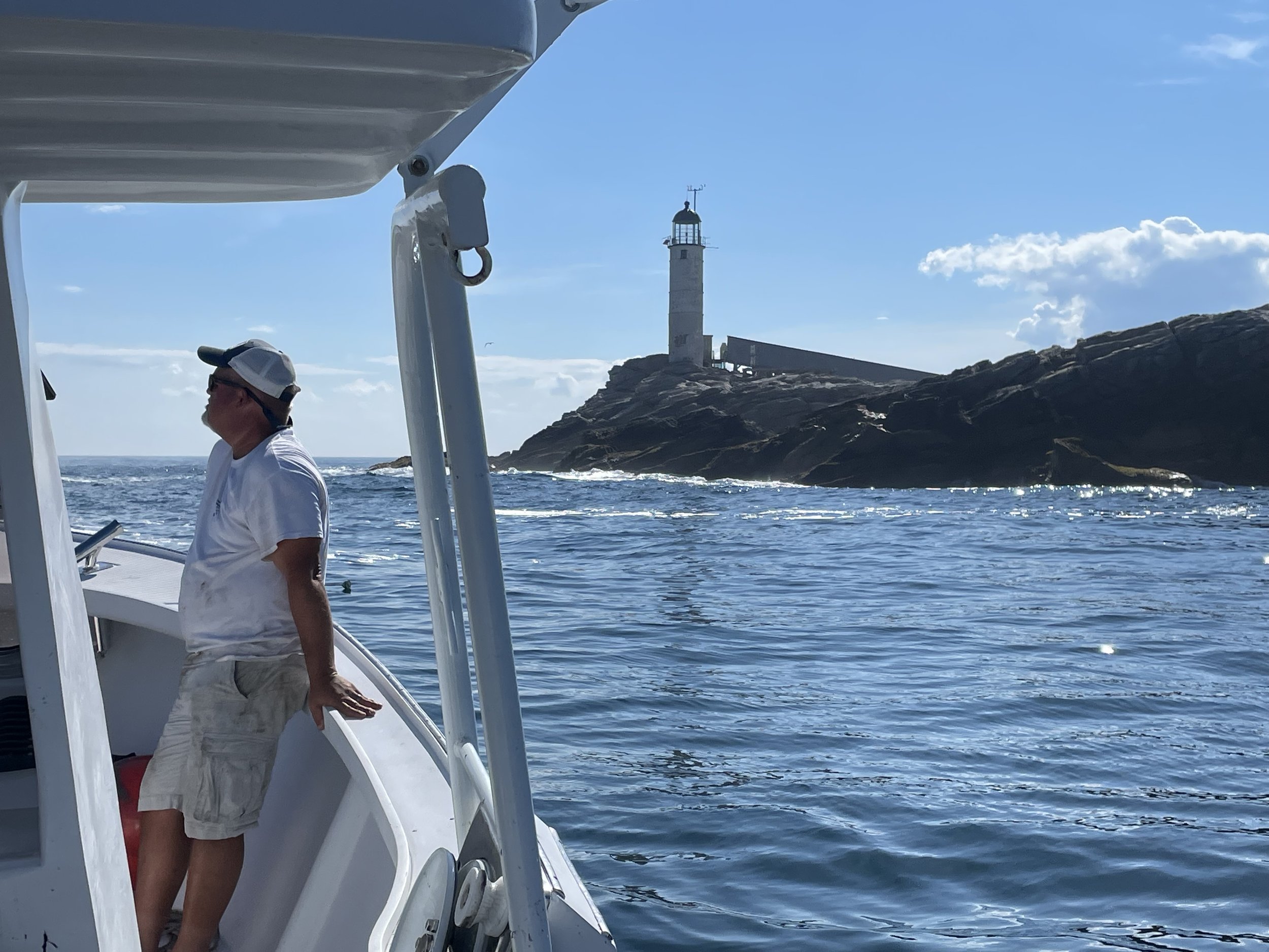A man leaning against the railing of a boat, looking toward a rocky coastline with a lighthouse on top, on a sunny day with calm water.
