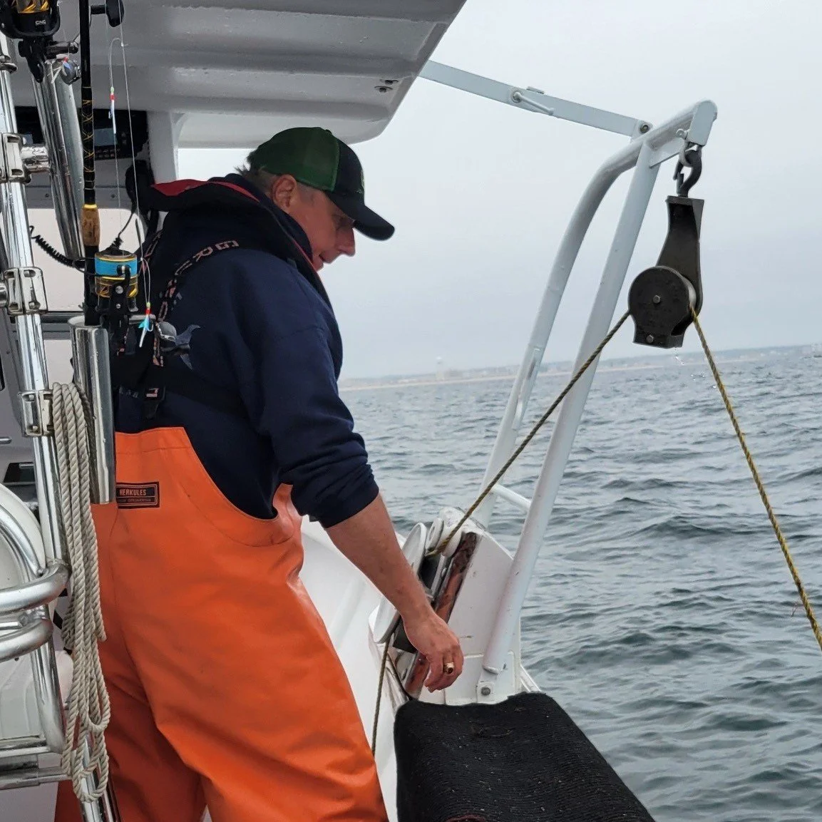 A man on a boat hauling lobster traps, with water and a distant shoreline in the background.