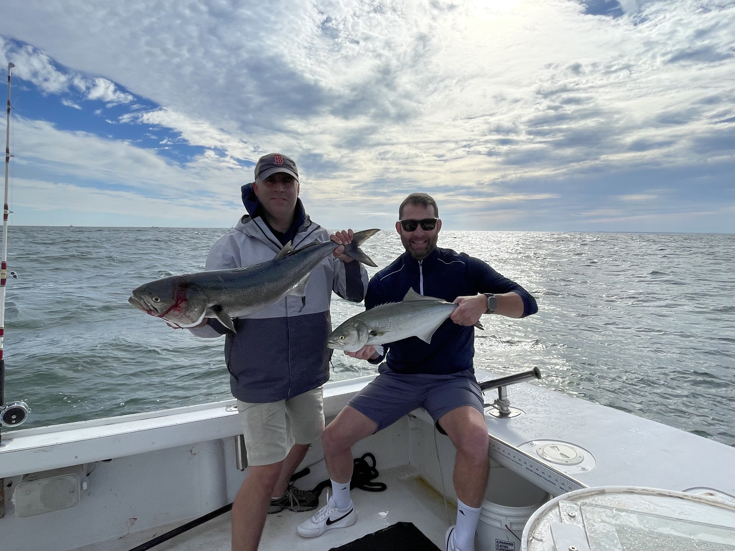 Two men on a boat holding large fish they caught while fishing in the ocean, with a partly cloudy sky in the background.