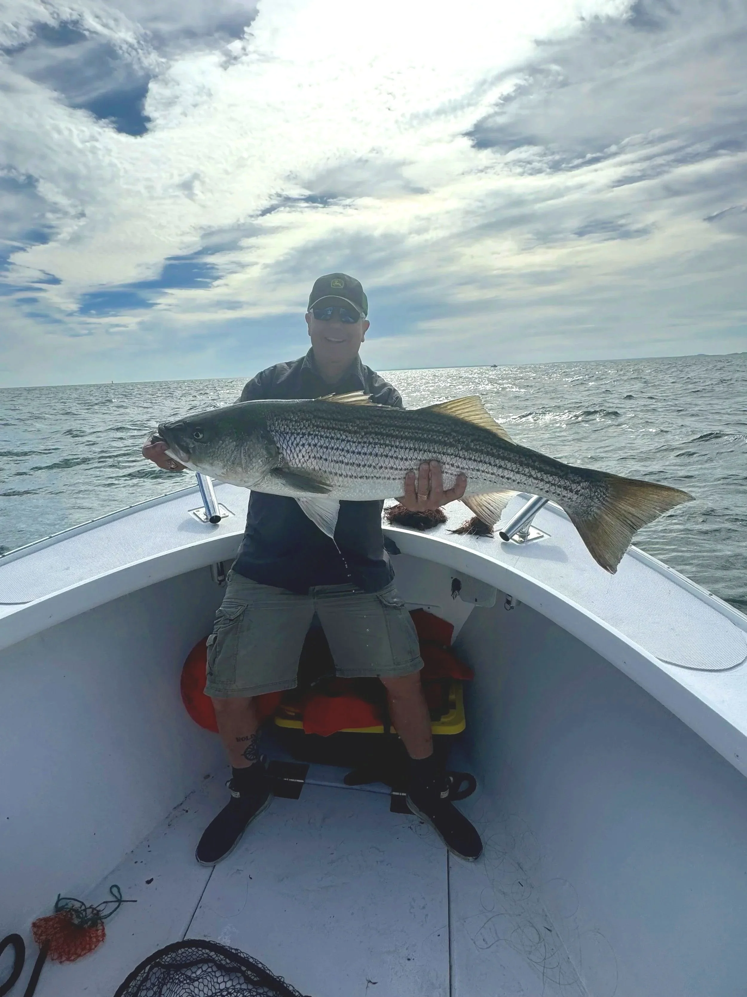 Man on a boat holding a large striped bass fish, wearing sunglasses and a black jacket, with the ocean and cloudy sky in the background.