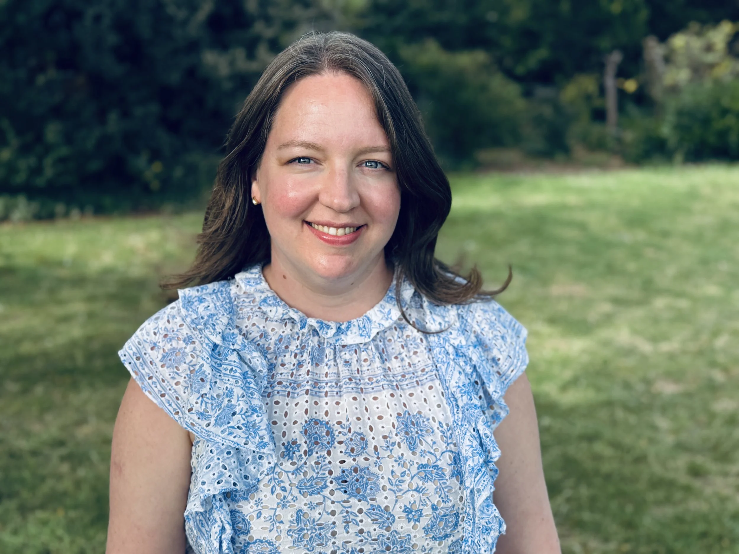 A smiling woman with shoulder-length brown hair and blue eyes standing outdoors on a grassy area with trees in the background, wearing a blue and white floral dress.