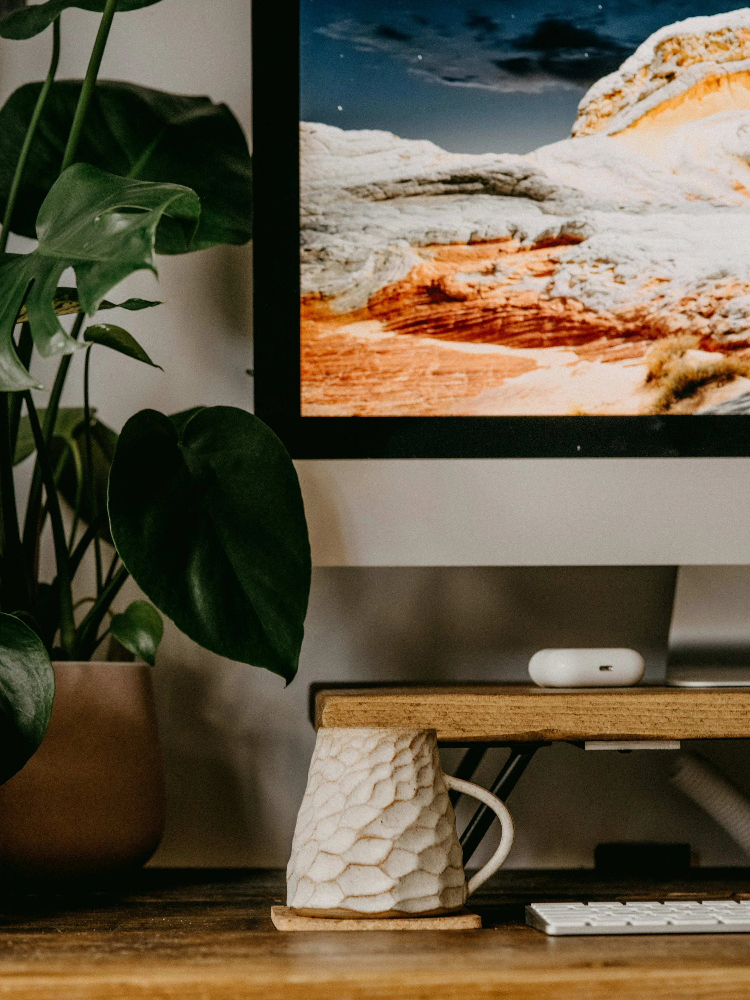 Close-up of a computer monitor displaying a mountain landscape, with a potted plant on the left and a textured white ceramic mug on a wooden surface below.
