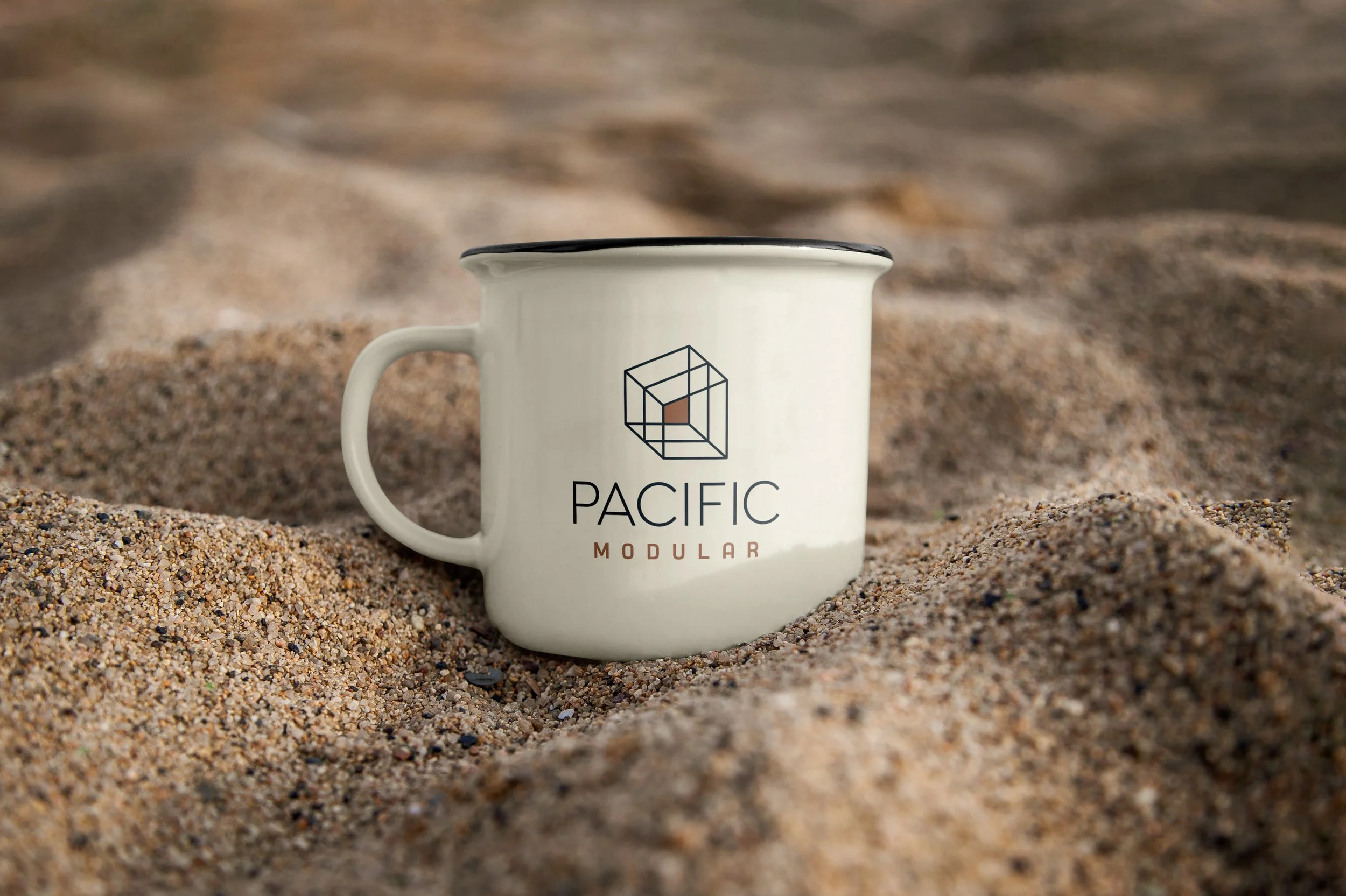 A white ceramic mug with the words 'Pacific Modular' and a geometric logo, placed on sandy terrain.