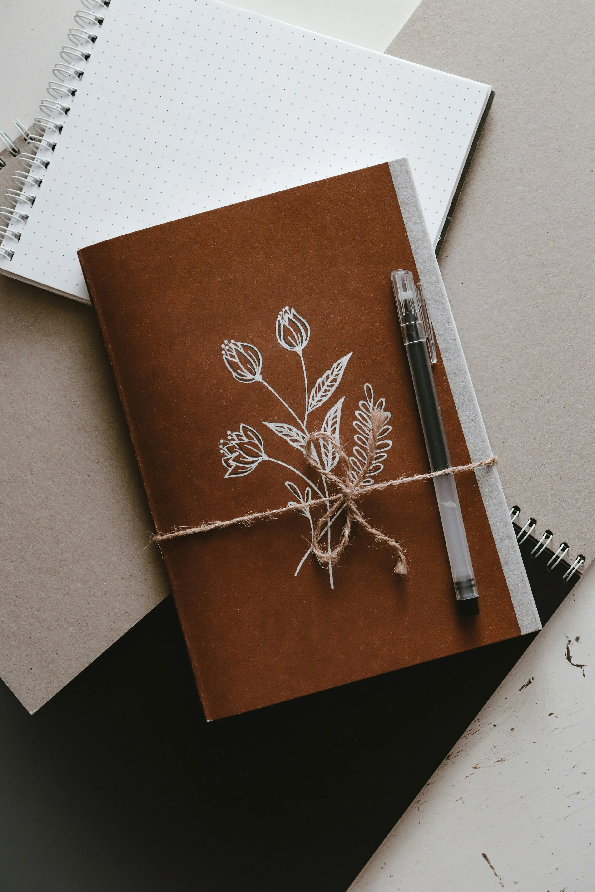 A brown notebook with a white floral drawing on cover, tied with twine, some open notebooks surrounding it, and a pen on top.