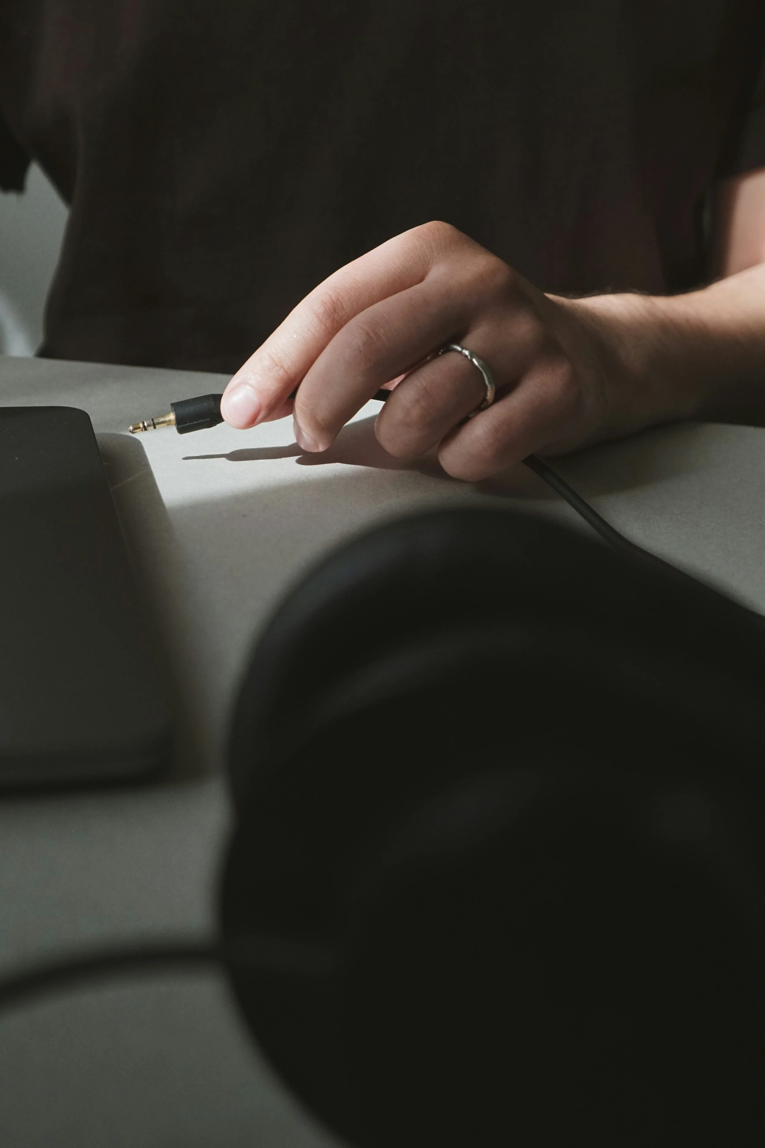 A person holding a black audio cable with a gold connector, ready to plug it in, on a gray surface with a smartphone and a pair of headphones nearby.