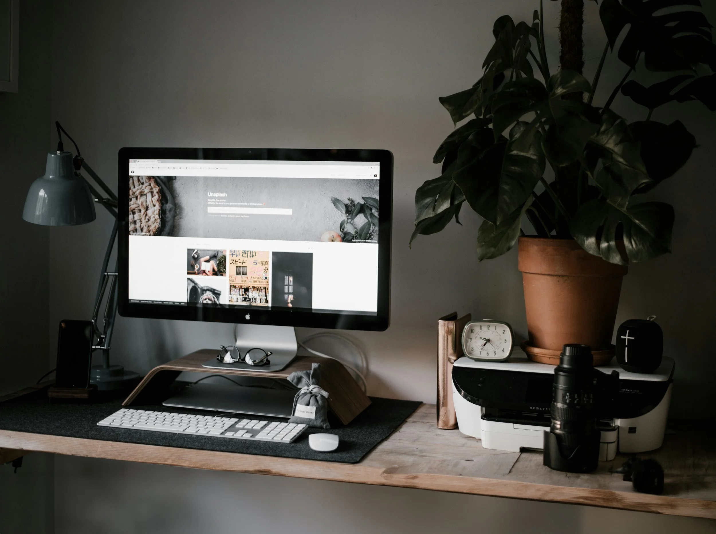 Home workspace with a computer monitor, keyboard, mouse, a desk lamp, glasses, potted plant, camera lens, clock, books, printer, and other small items on a wooden desk.
