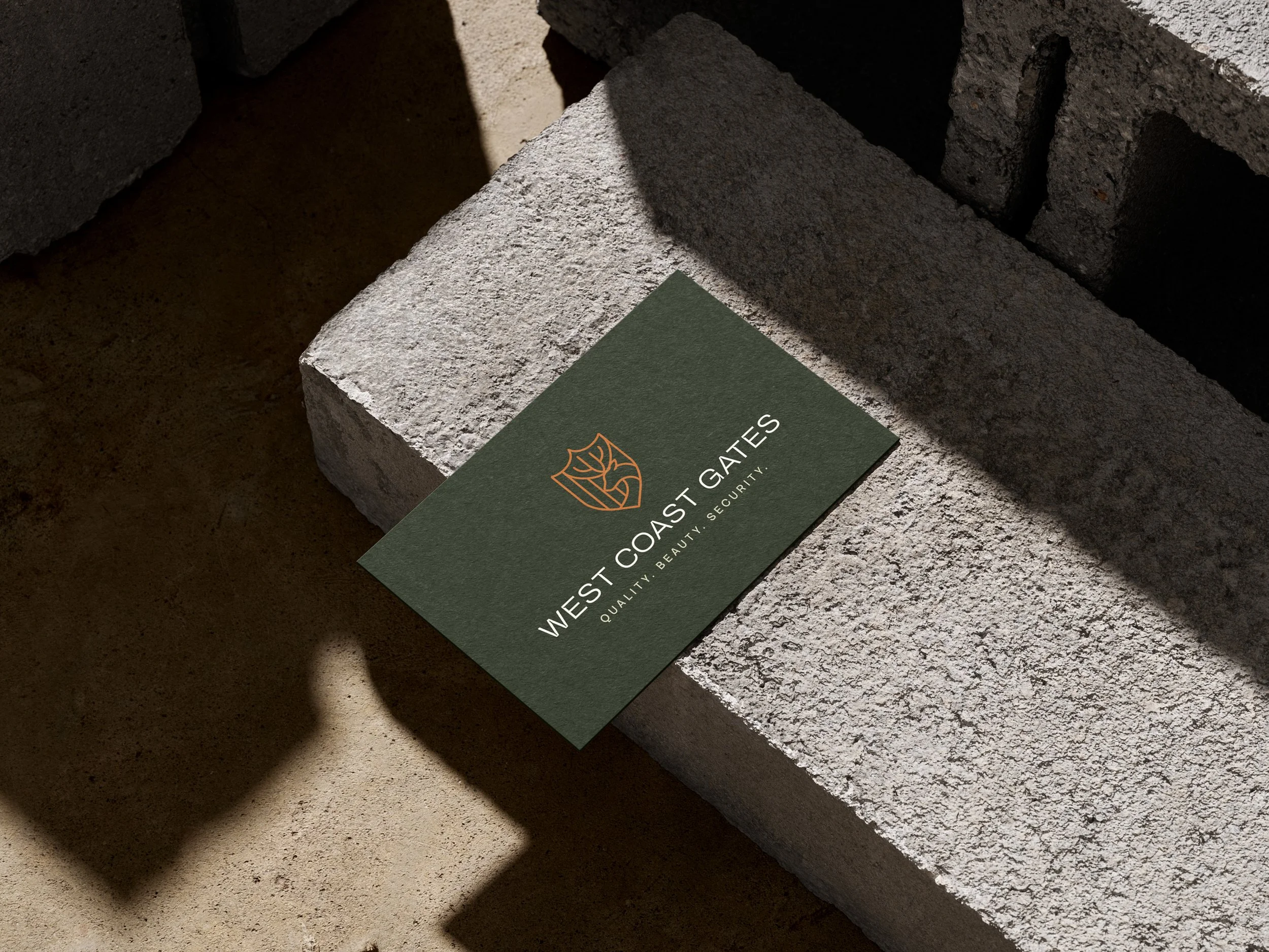 Business card for West Coast Gates with logo, black background and white text, on a concrete block with shadows.