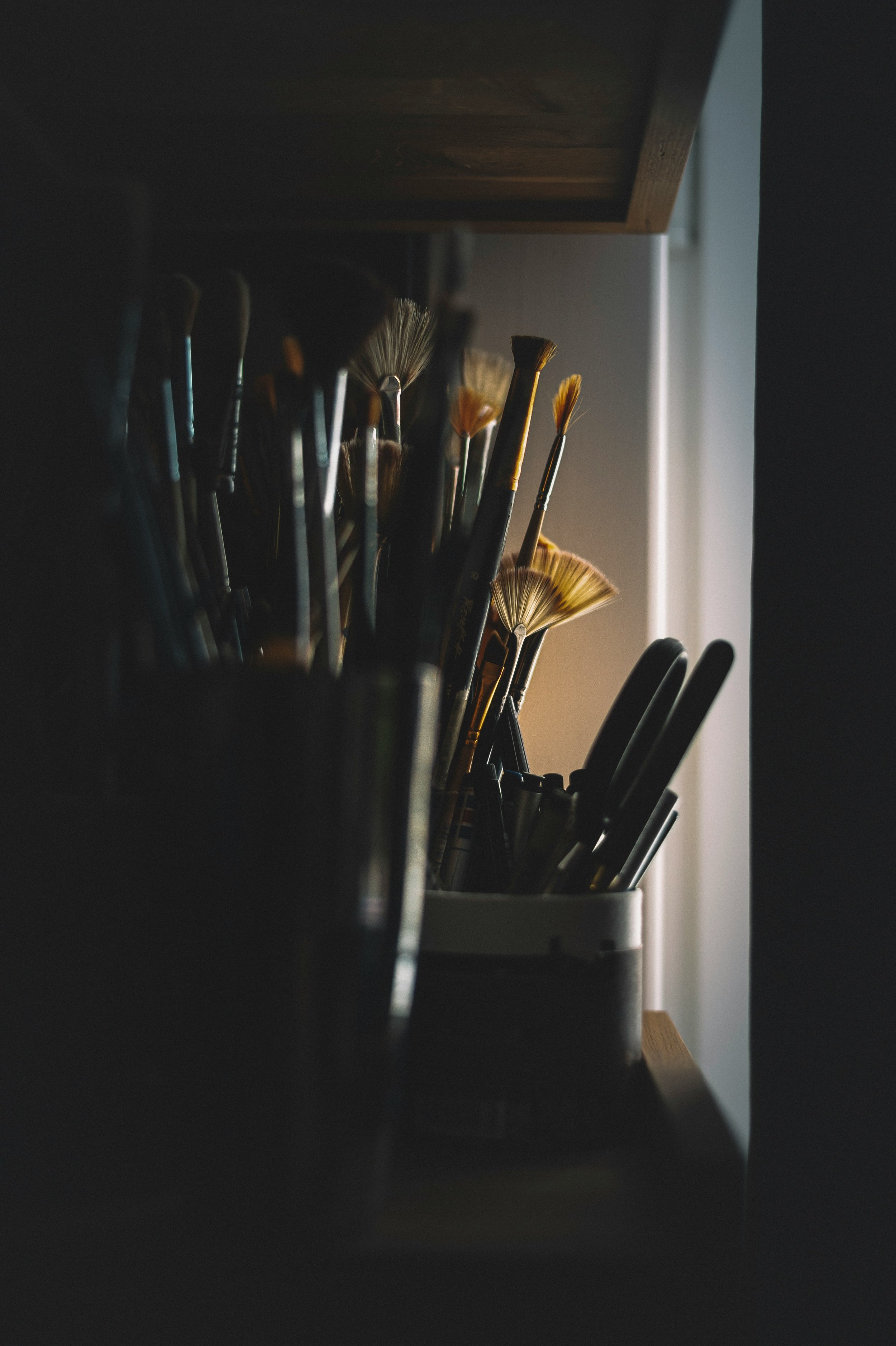 Paintbrushes in a container illuminated by natural light in a dim room.