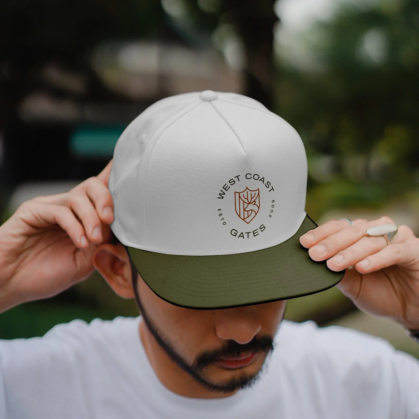 A person wearing a white and green cap with a logo that says 'West Coast Gates,' holding the brim, outdoors with blurred greenery in the background.