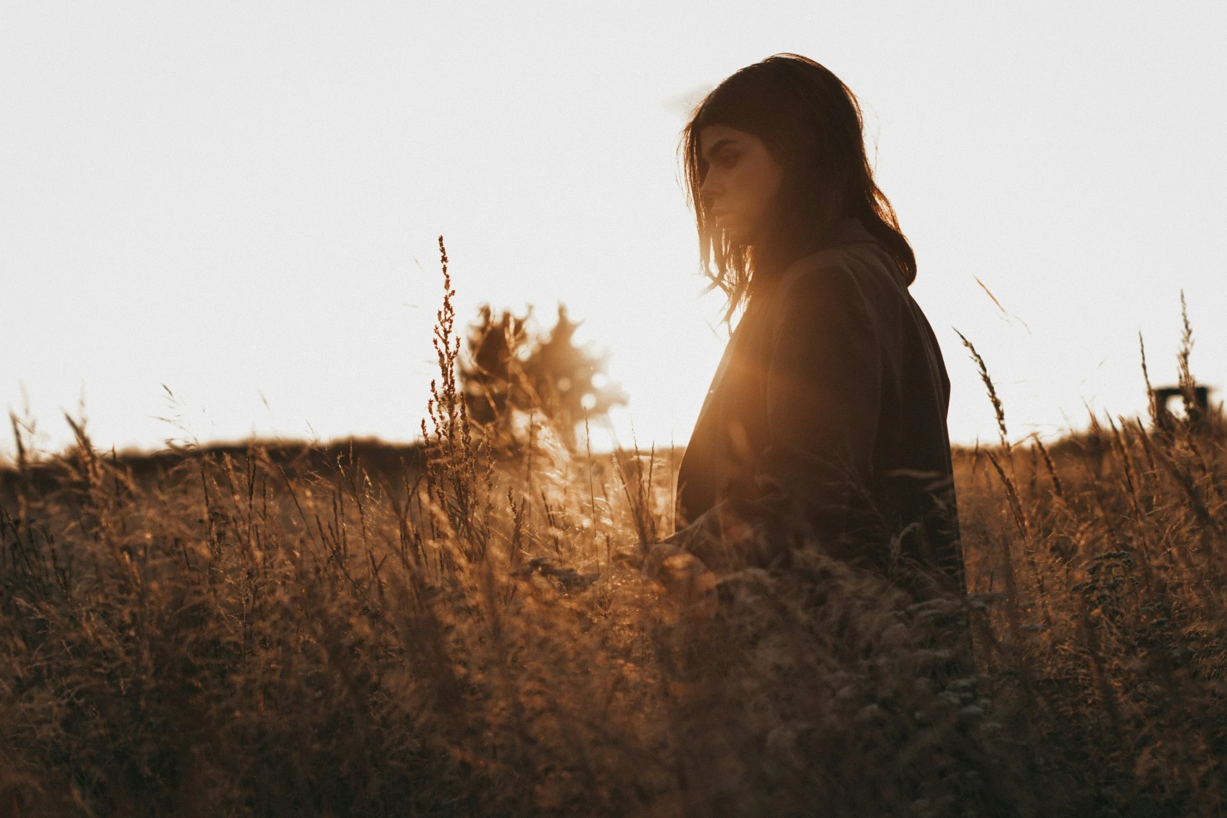 A woman with dark hair sitting in a field of tall grass during sunset, with the sun casting a warm glow around her.