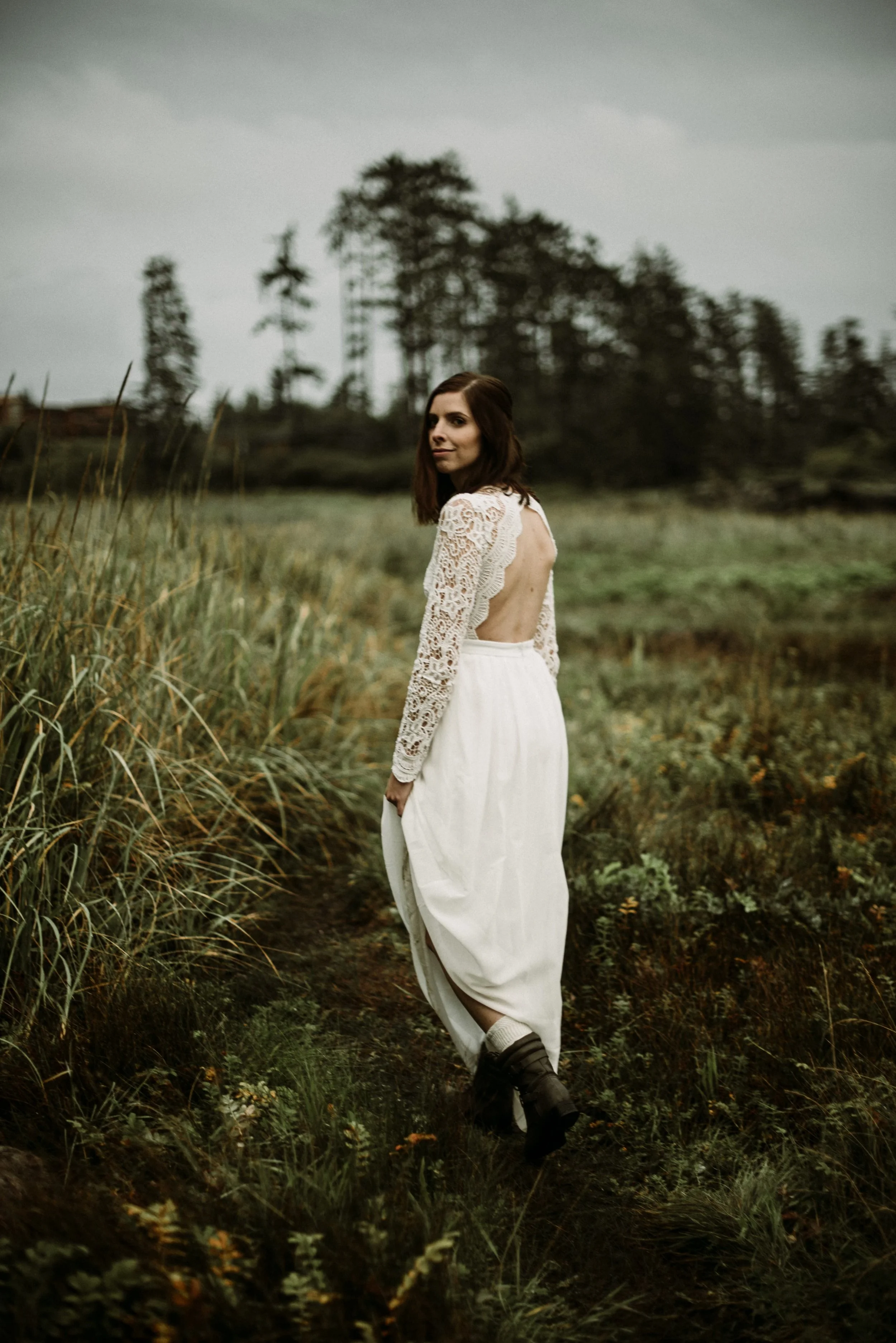 A woman in a white lace top and long white skirt standing outdoors on a grassy trail, with trees and an overcast sky in the background.