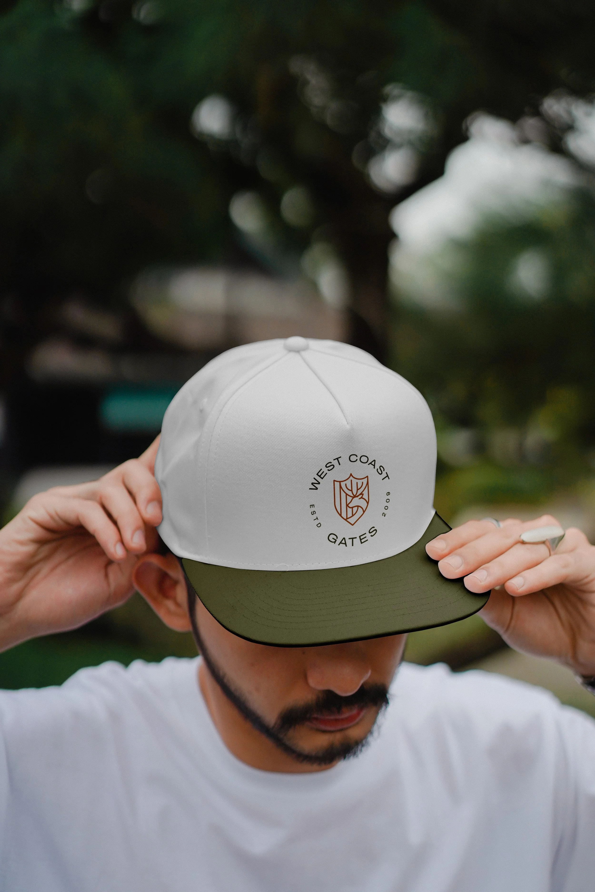 A man wearing a white t-shirt and a white and olive green cap with a logo that says 'West Coast Gates' and features a crest with a leaf design, outdoors with blurred trees in the background.