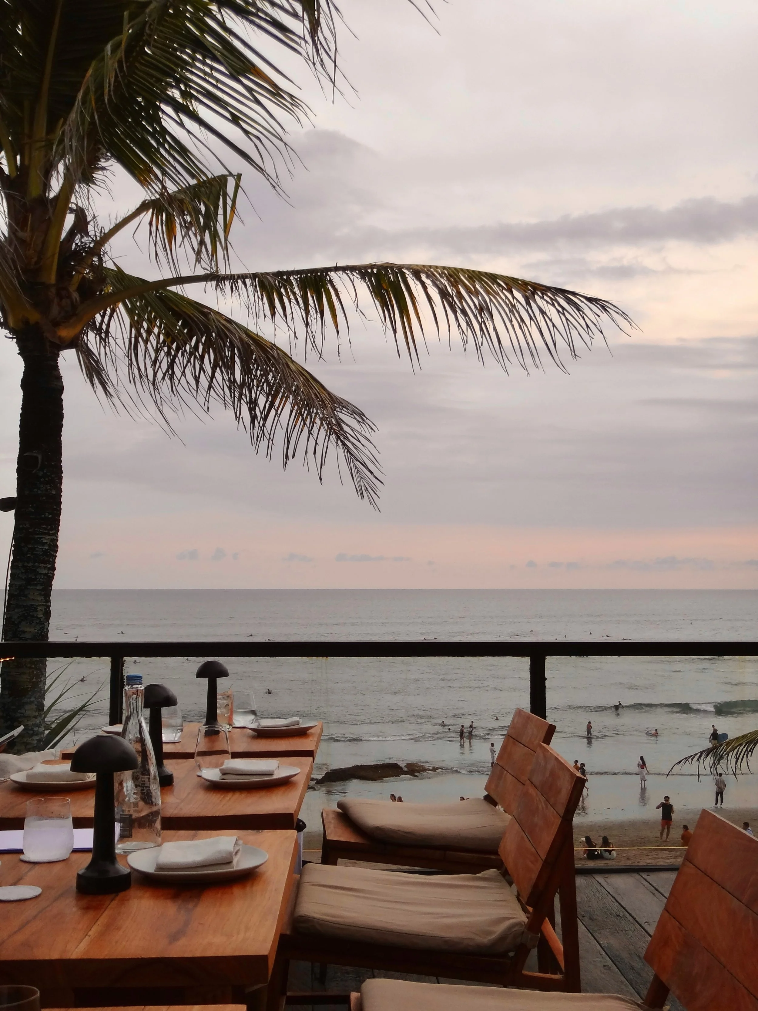 A beachside restaurant with wooden tables and chairs overlooking the ocean, palm trees, and people swimming and walking on the beach during sunset.