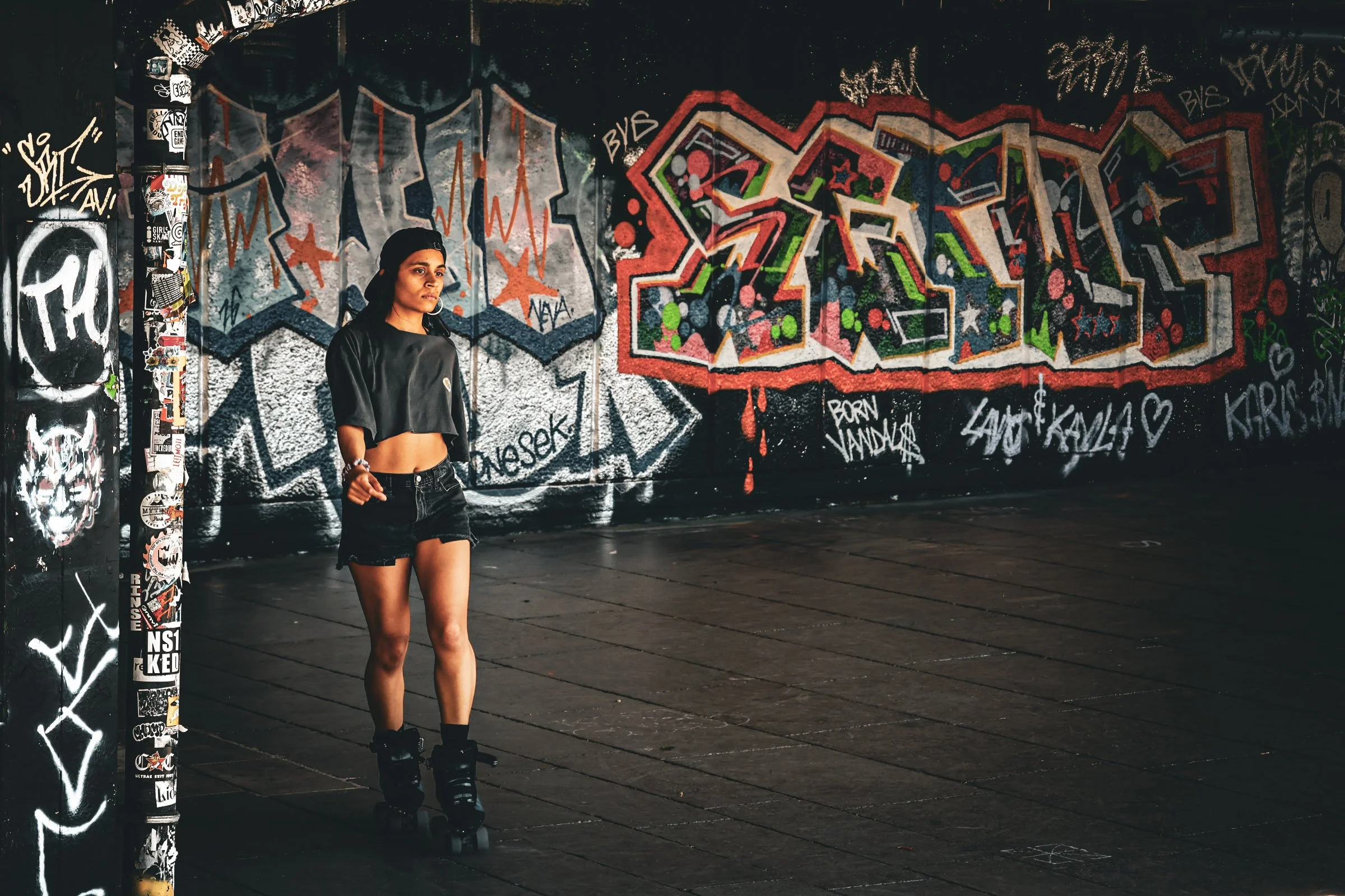 A woman in black roller skates, black shirt, and shorts stands in front of a graffiti-covered wall, with a sticker-covered pole to her left.