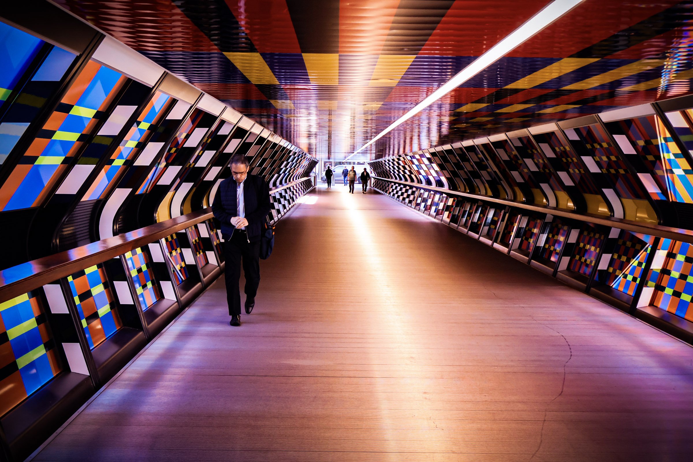 Colorful tunnel with diagonally patterned panels on the walls and ceiling, with a few people walking through, including a man in a suit looking at his phone.