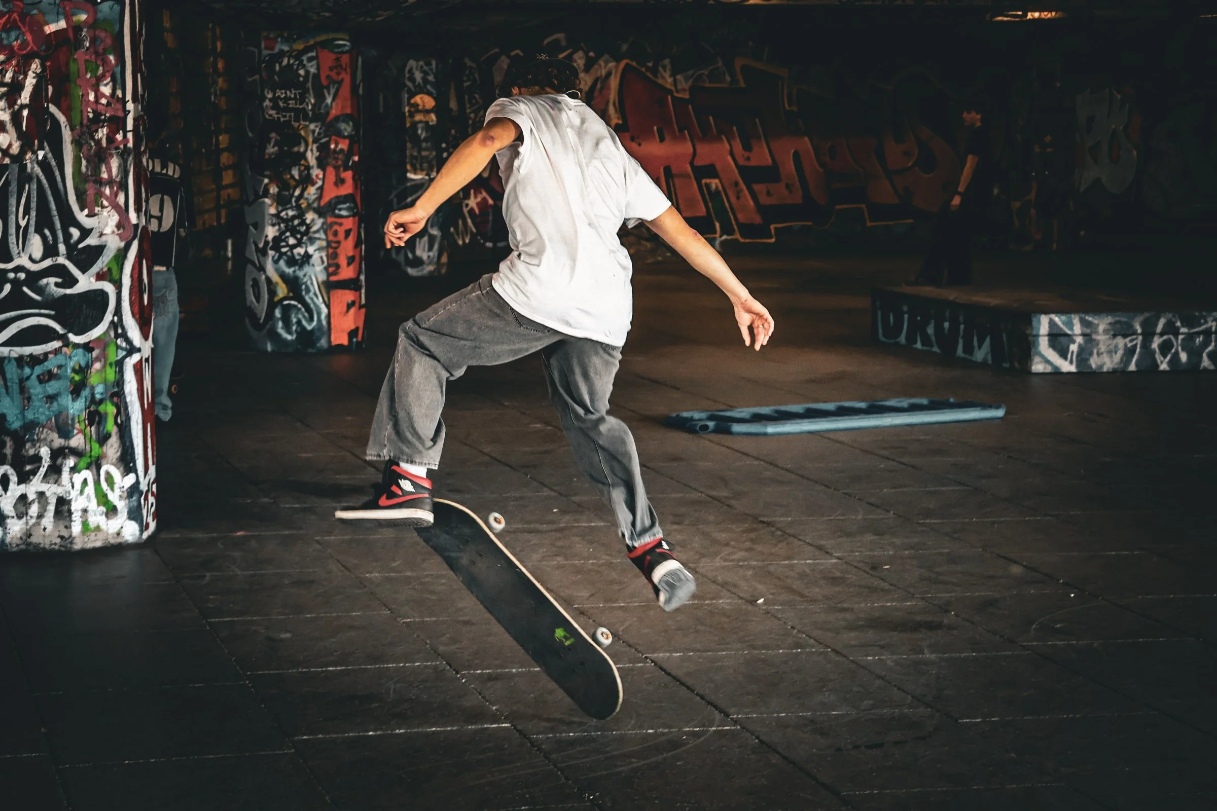 A person skateboarding indoors at a graffiti-covered skate park, wearing a white shirt, gray baggy pants, and black and red sneakers.