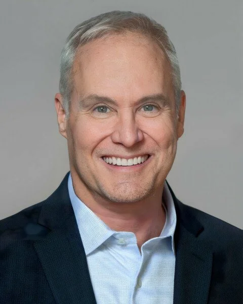 Thomas Downing, a smiling middle-aged man with gray hair, wearing a light blue shirt and a dark blazer, against a plain light gray background.