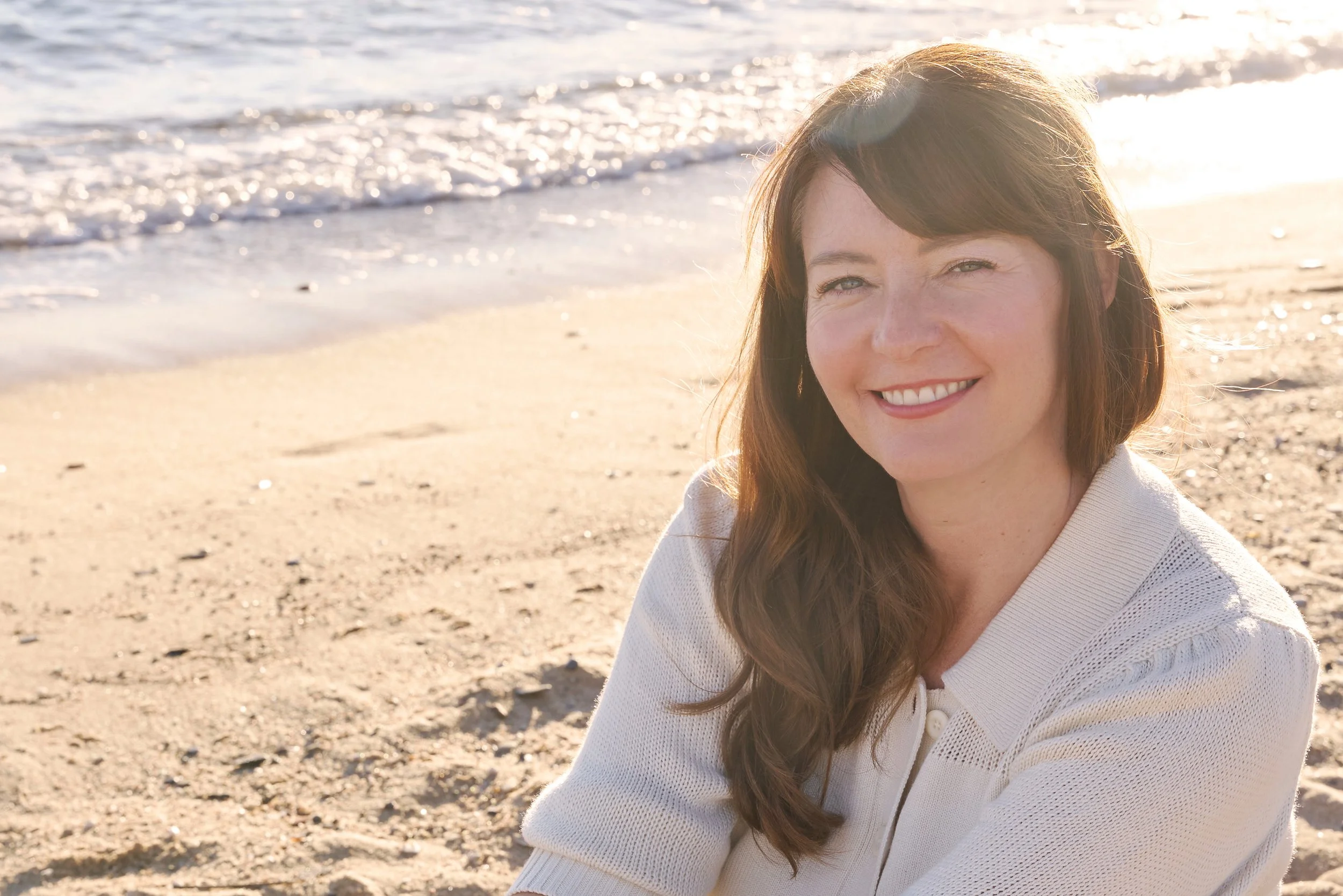 A woman with long brown hair smiling on a sandy beach near the ocean at sunset.