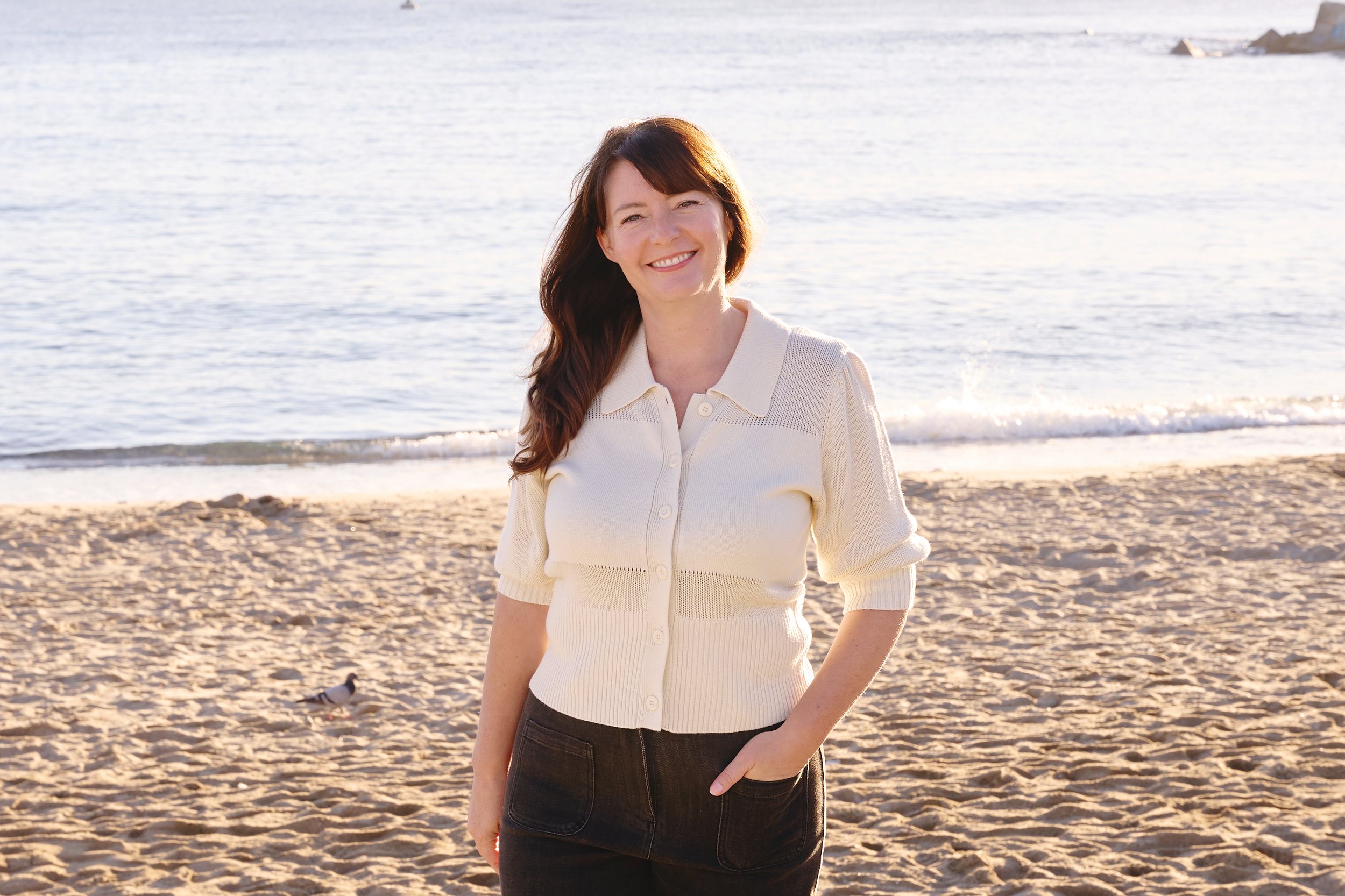 A woman with long brown hair wearing a cream-colored sweater and black pants standing on a sandy beach near the ocean, smiling at the camera during sunset.