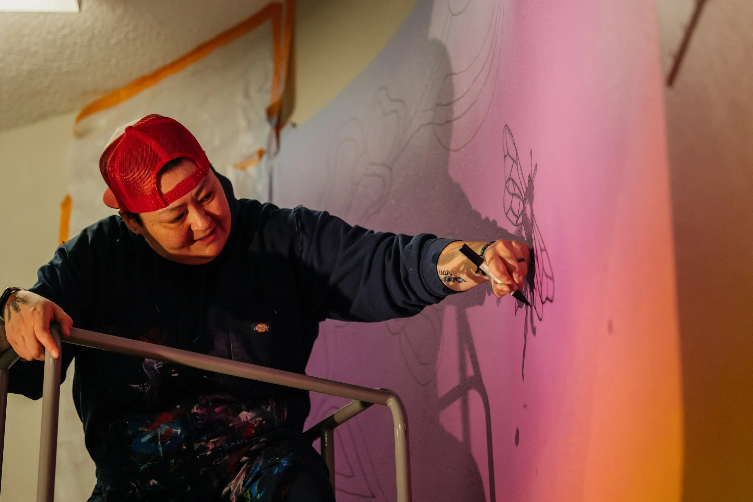 An artist wearing a red cap, black hoodie, and paint-splattered pants is on a scaffold drawing on a pink wall with black marker. The artist is focused on their work, and a shadow of their arm and marker is cast on the wall.