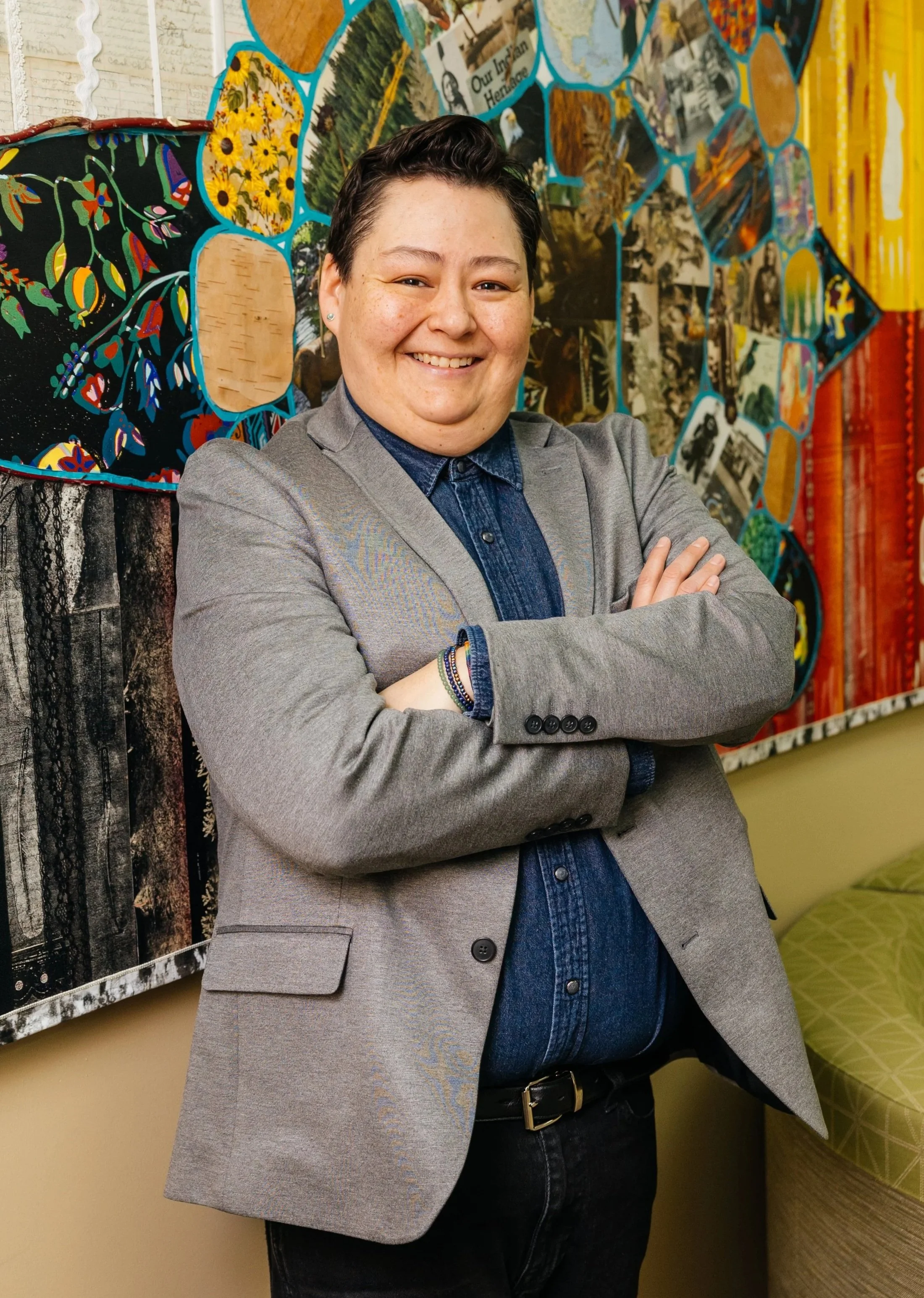 A smiling person with short dark hair, wearing a gray blazer over a denim shirt, standing with arms crossed in front of a colorful, patterned wall with various photos and artwork.
