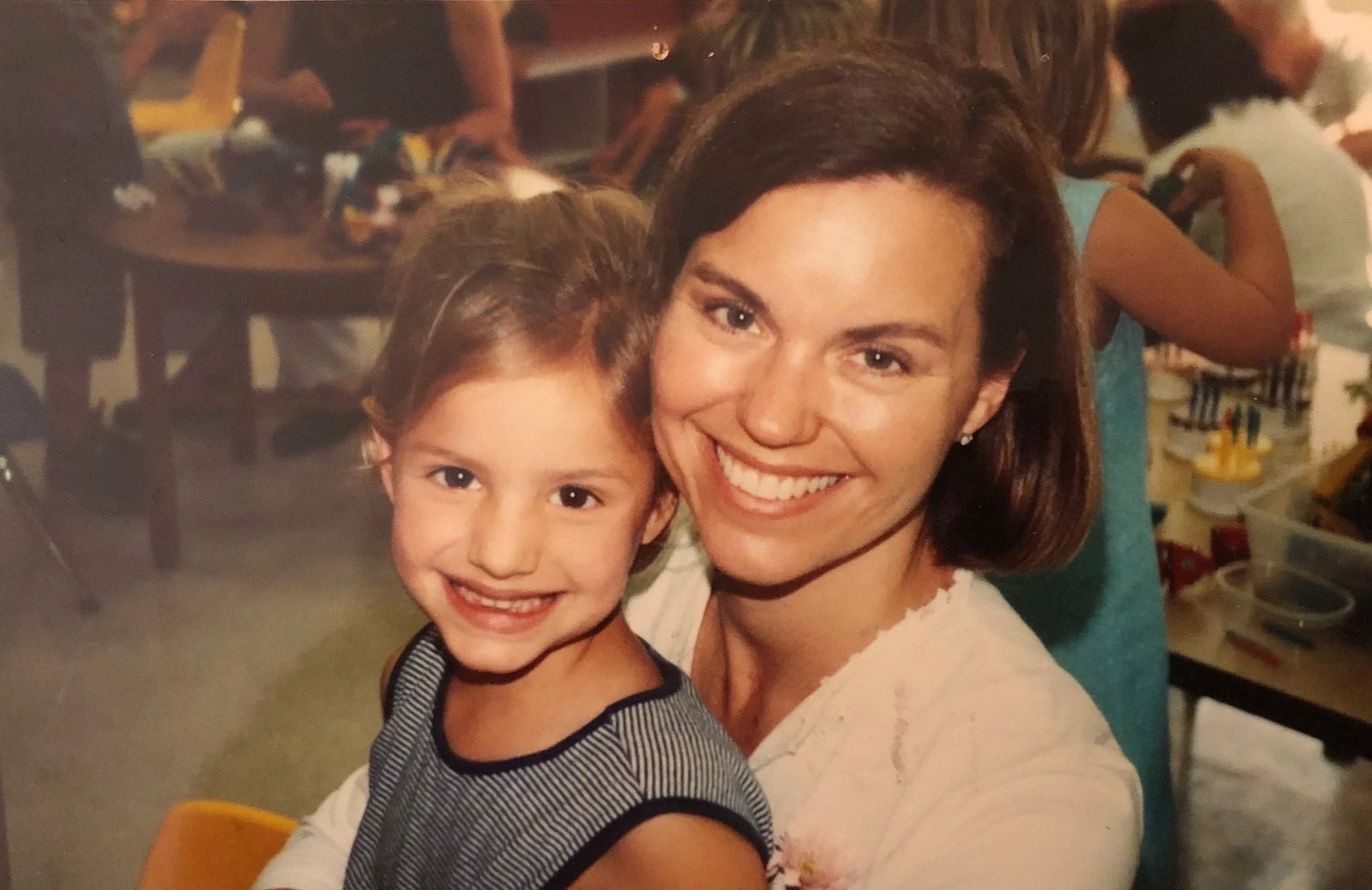 A woman and a young girl smiling and posing together in an indoor setting with a blurred background of people and tables.