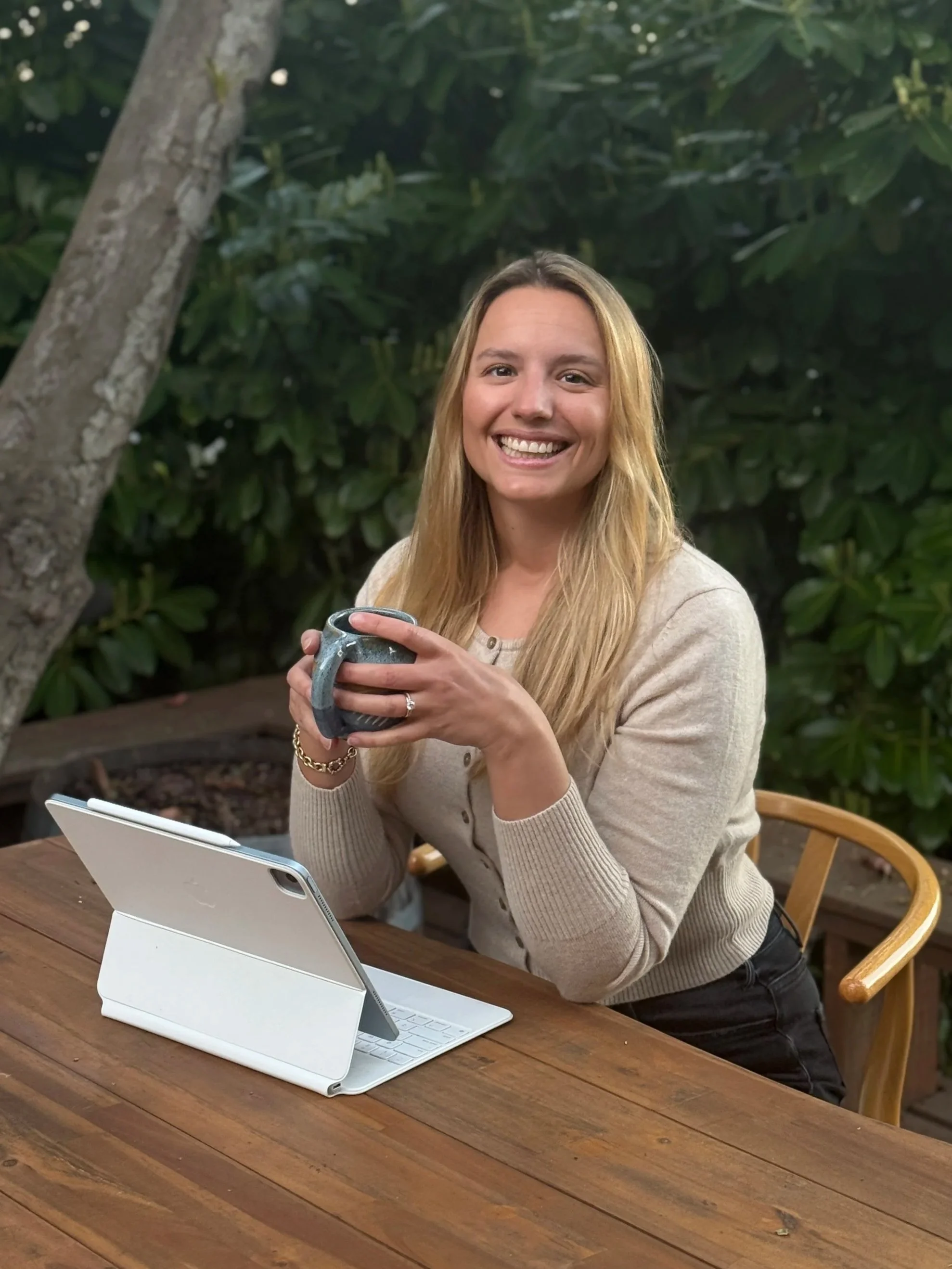 A woman sitting at a wooden table outdoors, holding a dark gray mug, smiling, with a tablet computer in a white case on the table, surrounded by greenery.