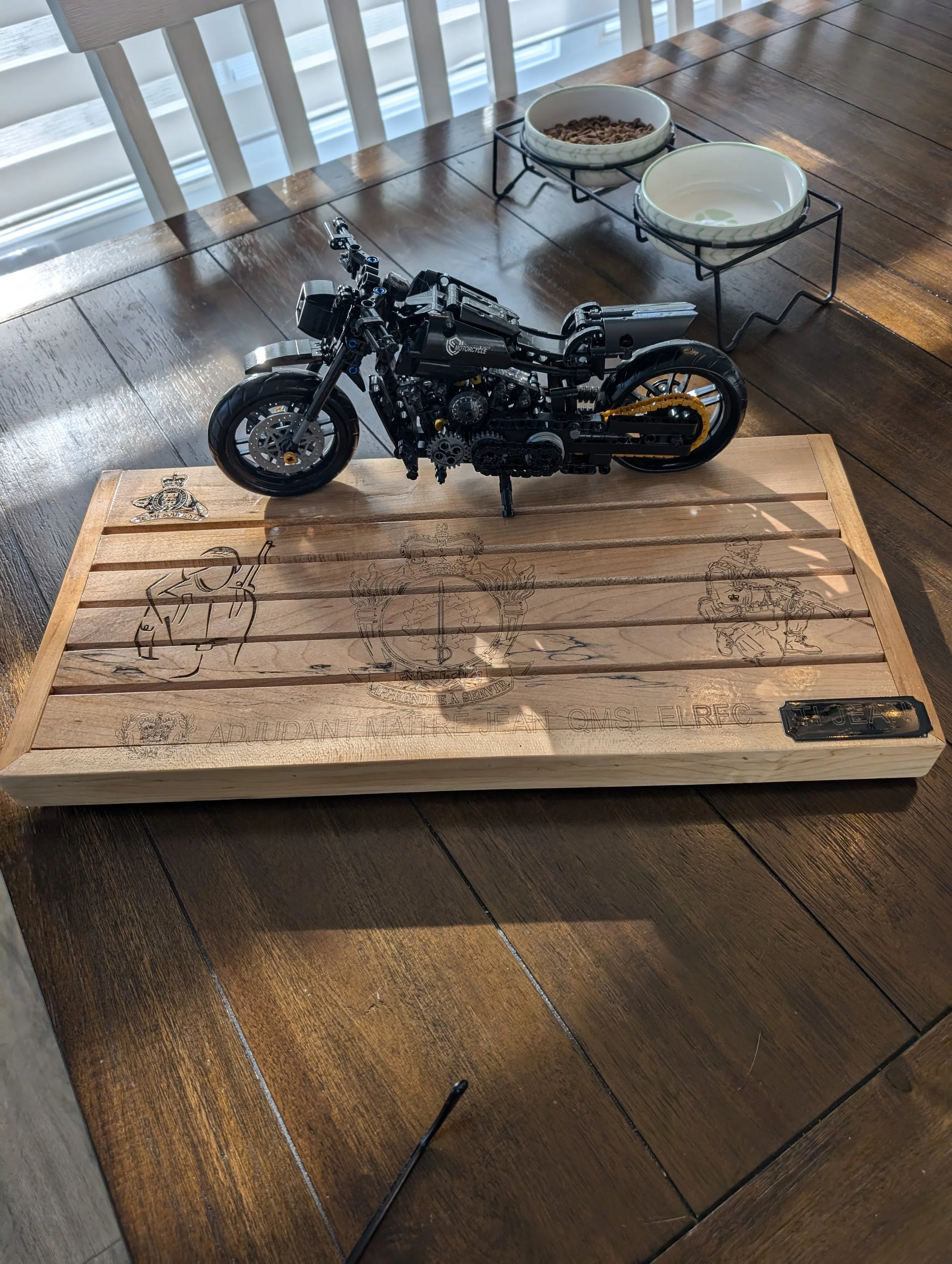 Une maquette de moto construite avec des pièces en plastique noir, placée sur une table en bois. Deux bols en céramique, un contenant pour la nourriture pour animal et une grille en métal sont visibles en arrière-plan, posés sur la même table.