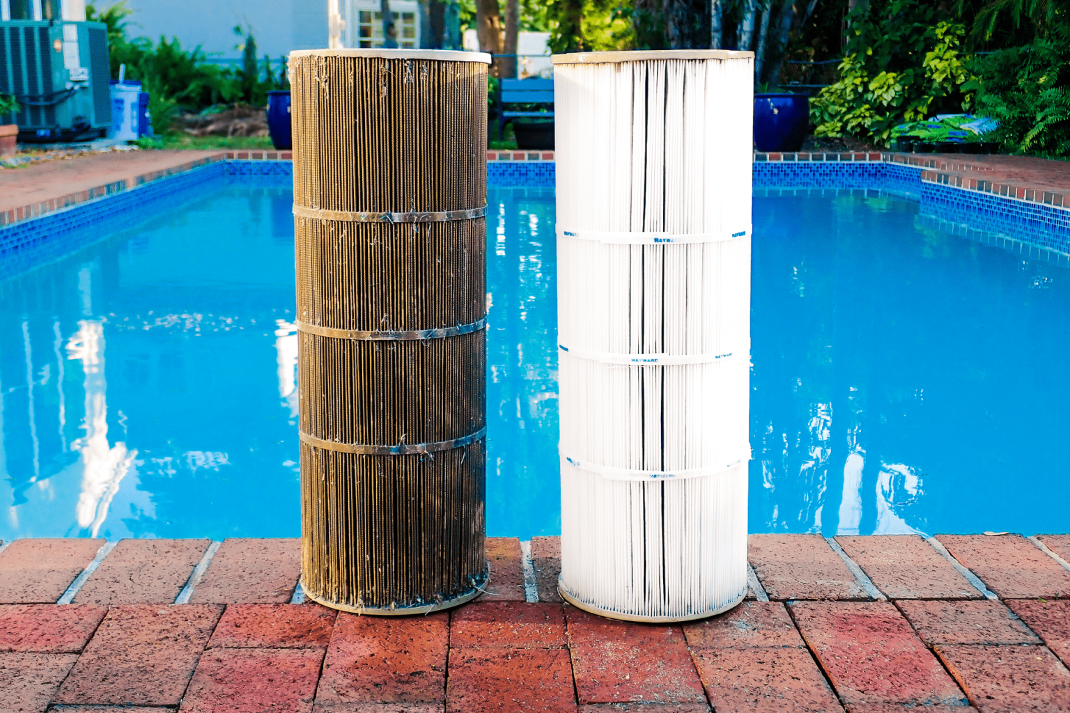 Dirty pool filter on the left and clean pool filter on the right standing on brick pool deck beside a swimming pool with blue water, surrounded by green plants and outdoor furniture.