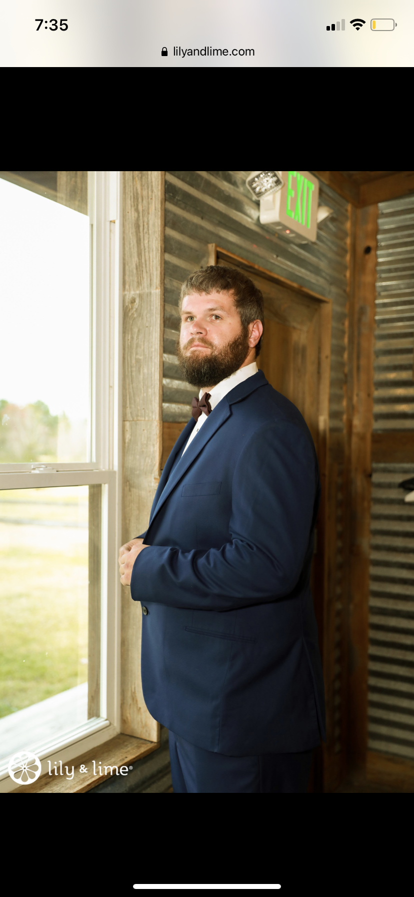 A man with a beard wearing a navy suit, white shirt, and bow tie standing indoors near a window with a rustic wooden wall behind him.
