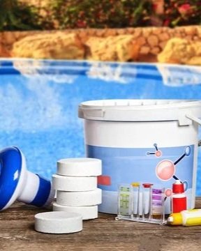 Poolside scene with a bucket of pool chemicals, including test tubes and tablets, near a swimming pool.