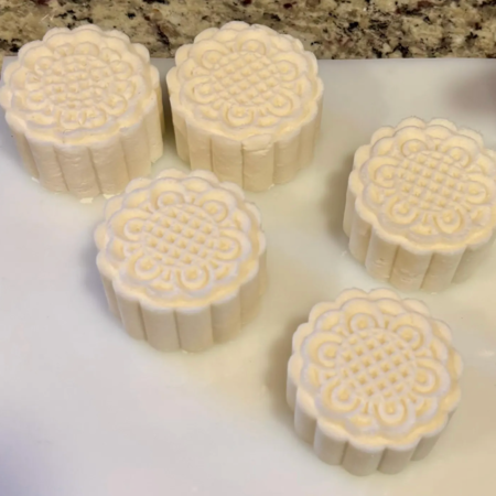 Six white mooncakes with intricate patterns on top, arranged on a white surface with a granite countertop in the background.