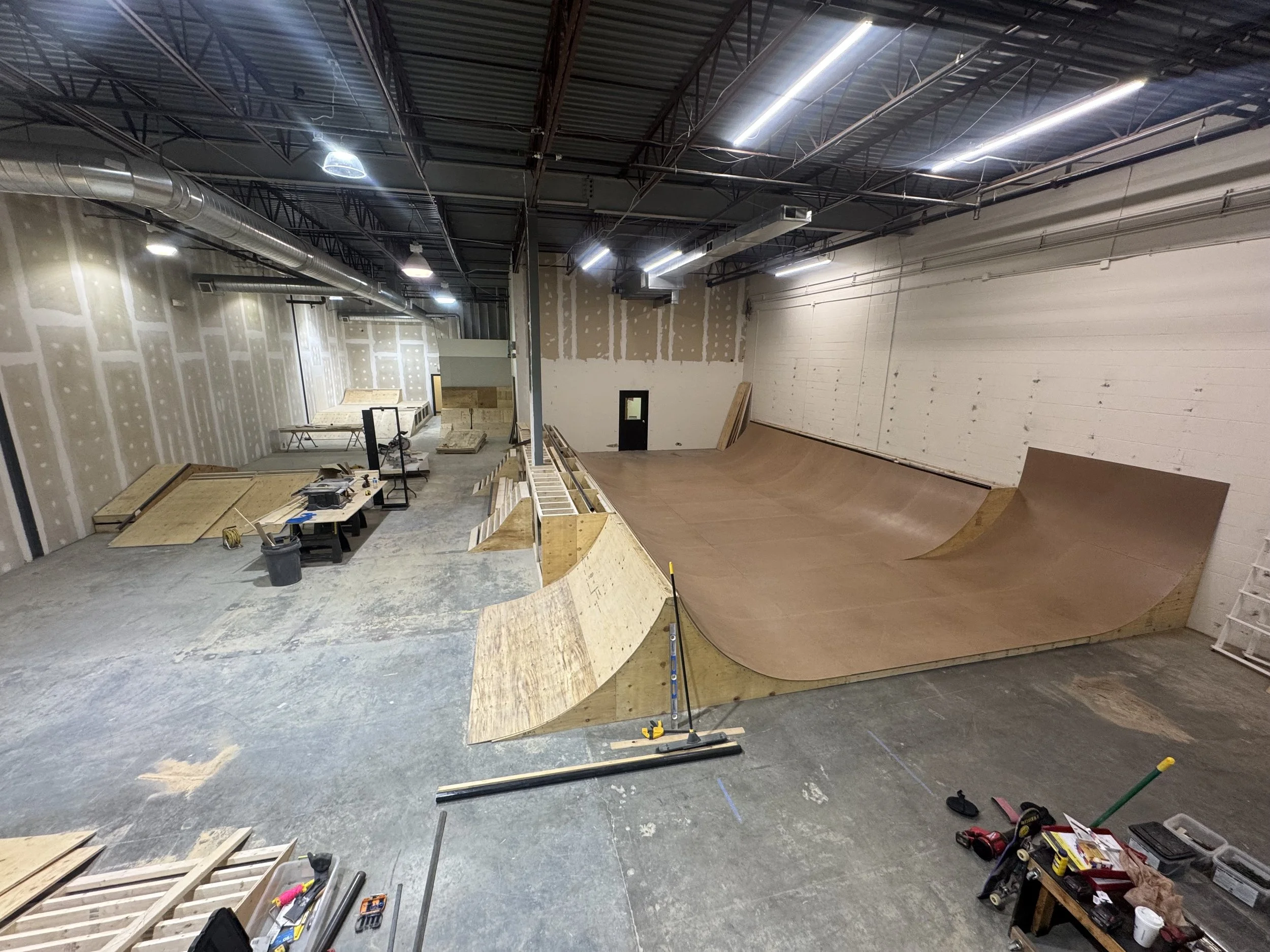 An indoor skatepark with wooden ramps and a half-pipe in progress. Construction tools and materials are scattered around.