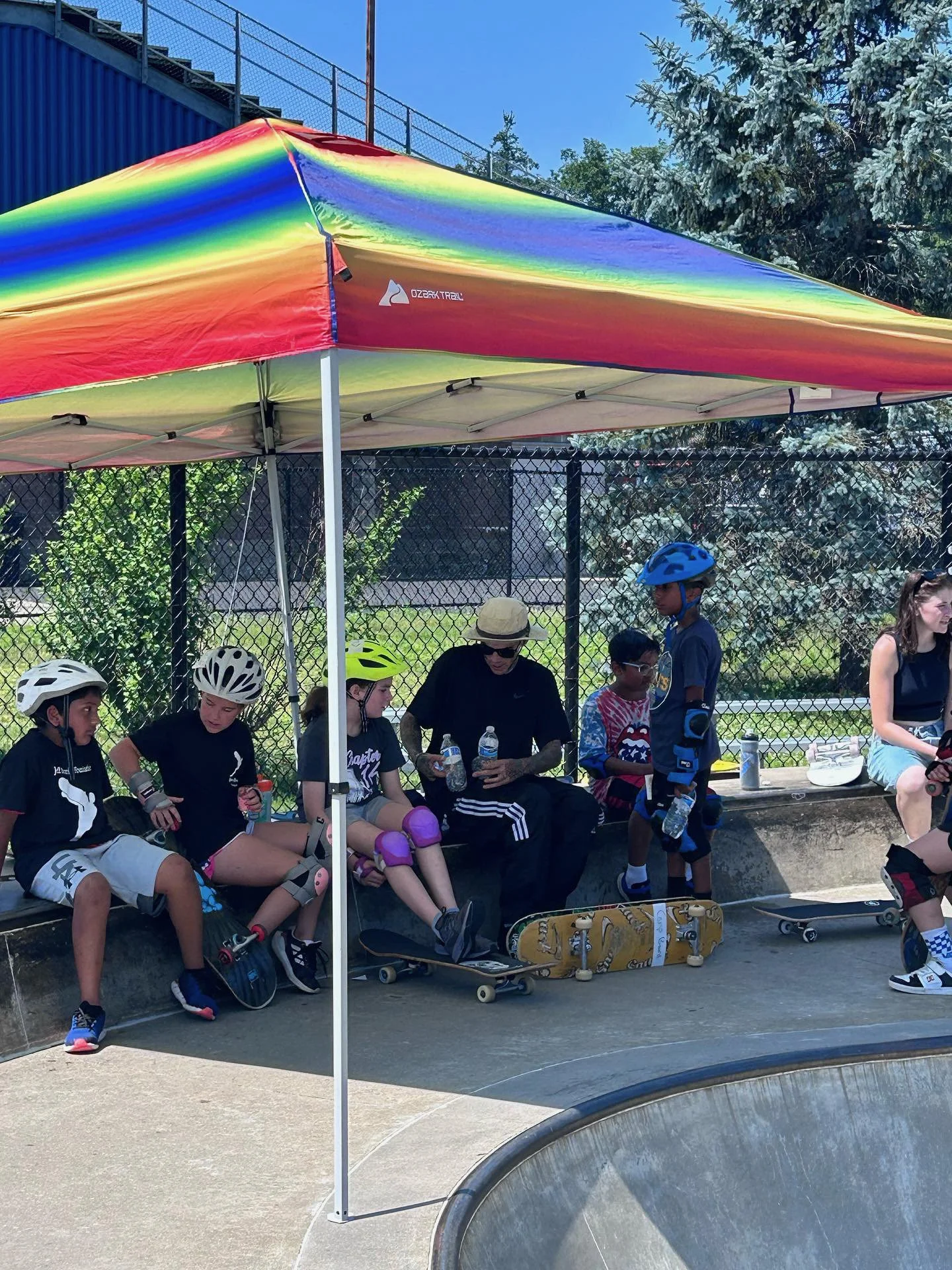 Children and an adult sitting on a bench under a rainbow-colored canopy at a skatepark, some wearing helmets and knee pads, with skateboards at their feet.