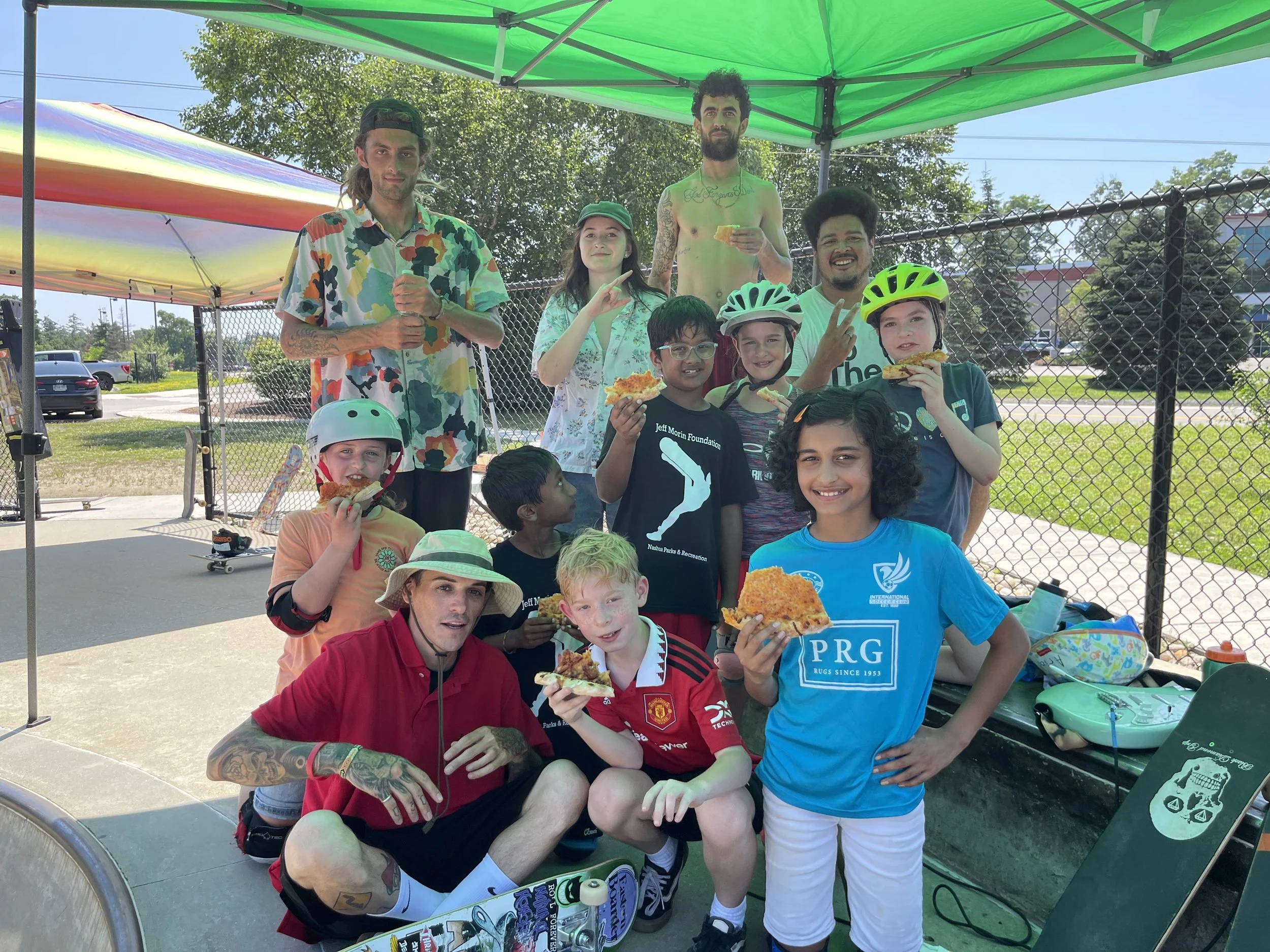 Group of children and adults at a skate park enjoying pizza, some wearing helmets, under a green canopy on a sunny day.