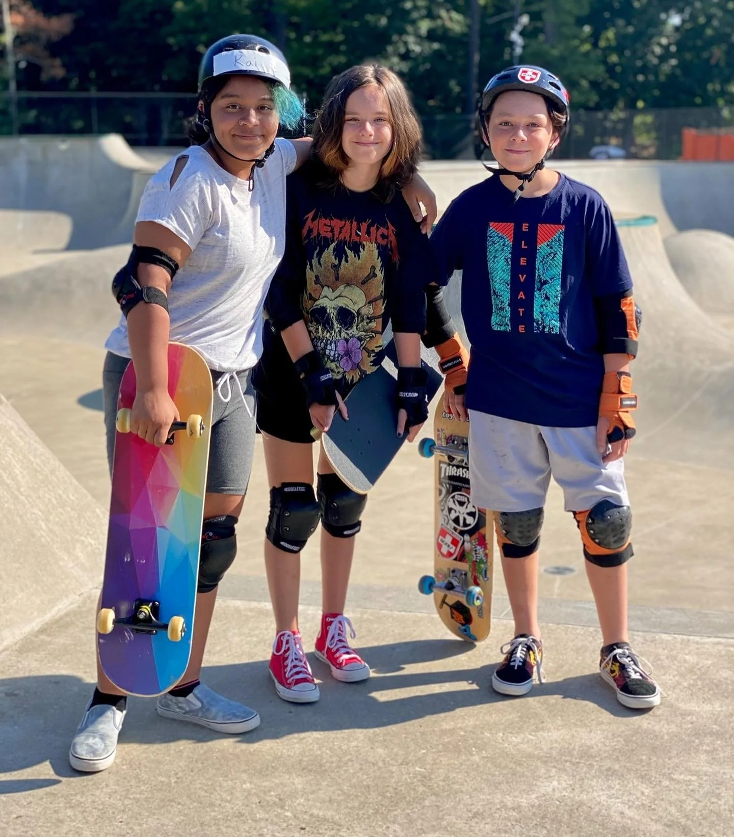 Three kids in skateboarding gear and helmets at the skate park, holding skateboards.