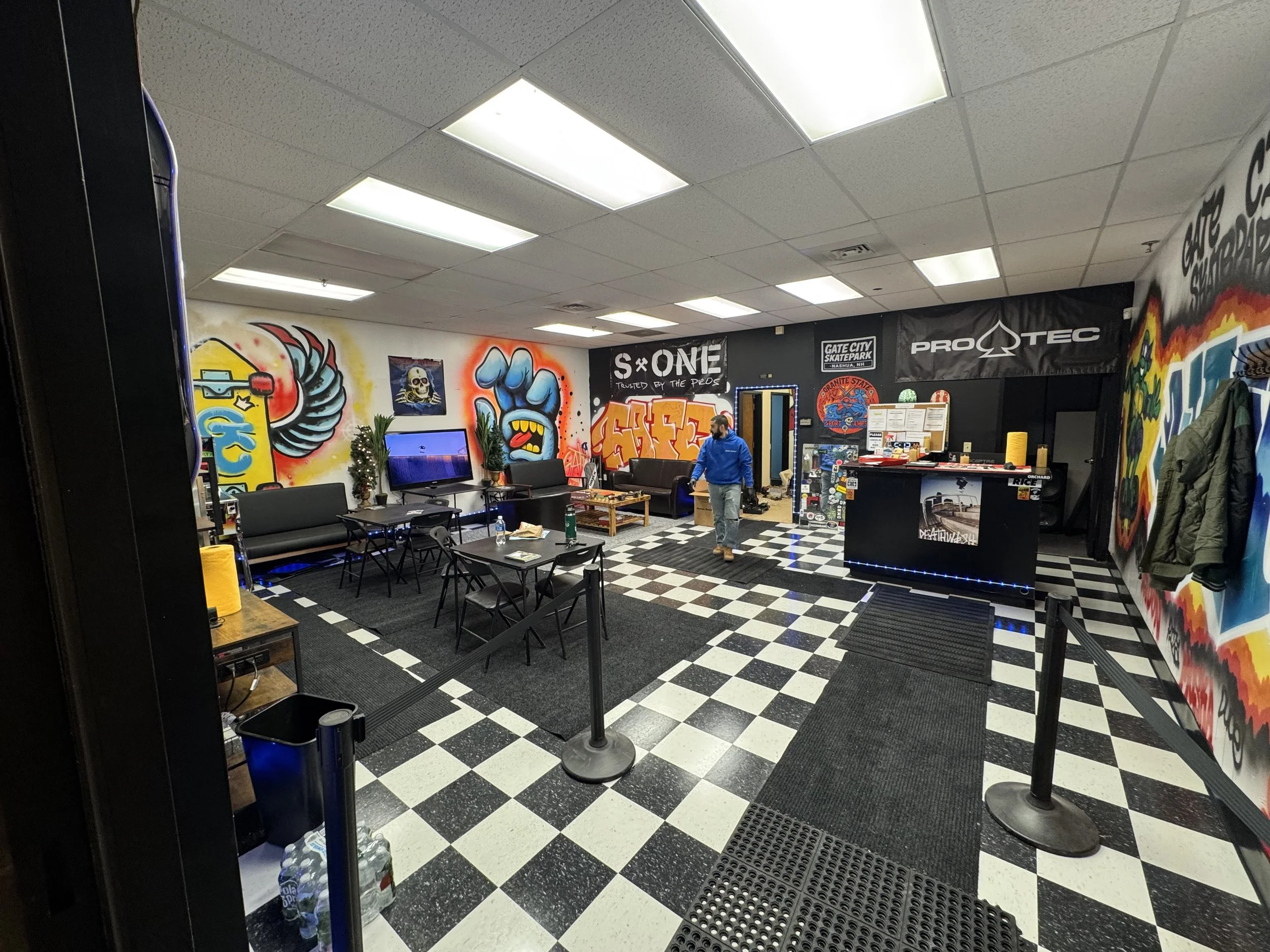 Indoor skate shop with black and white checkered floor, seated area with black chairs and tables, graffiti art on walls, counter at the back with skateboards and merchandise, person in blue jacket walking inside.