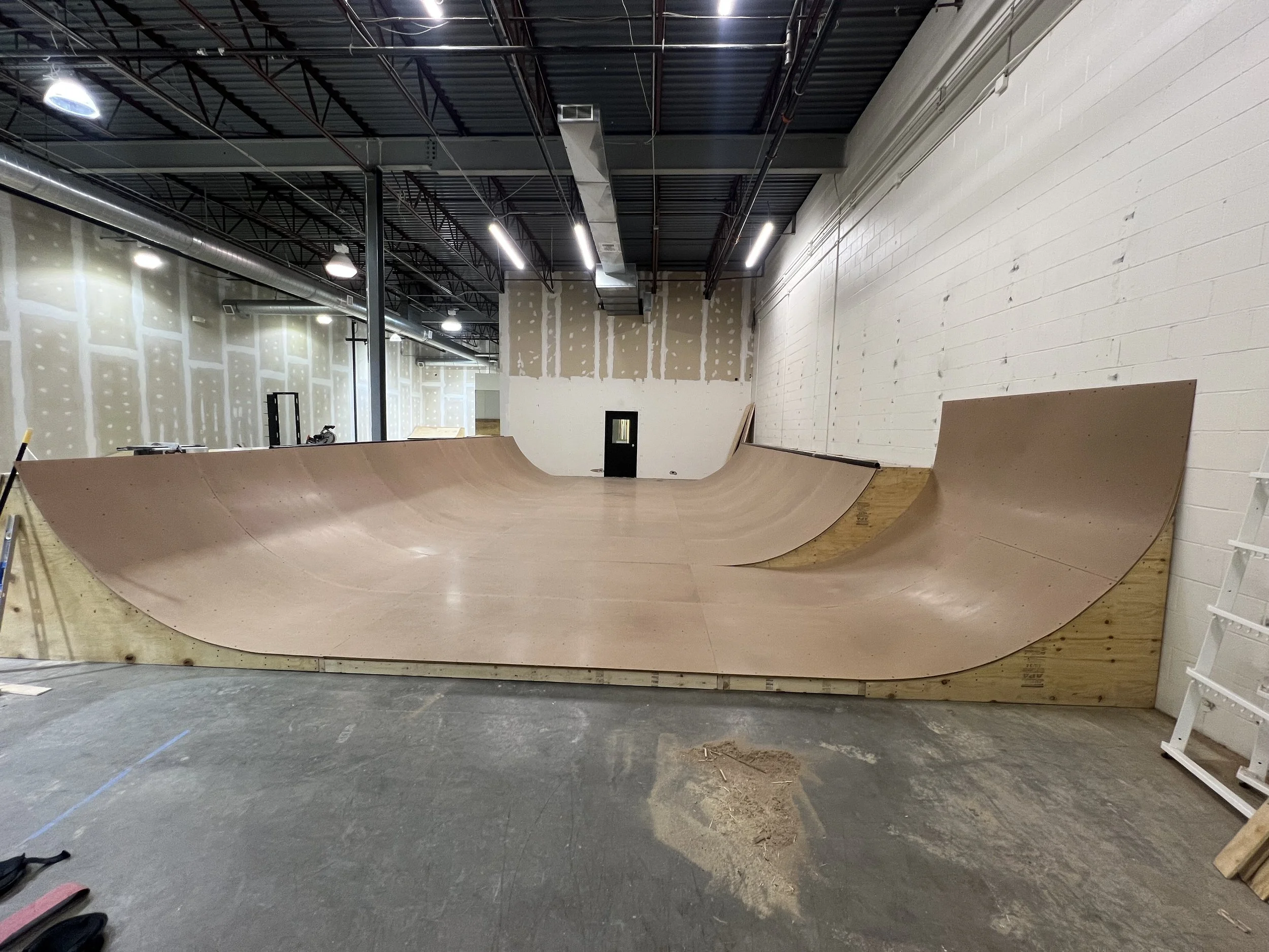 Indoor skatepark with unfinished wooden ramp and half-pipe in construction, exposed ceiling pipes and lighting, and white brick walls.