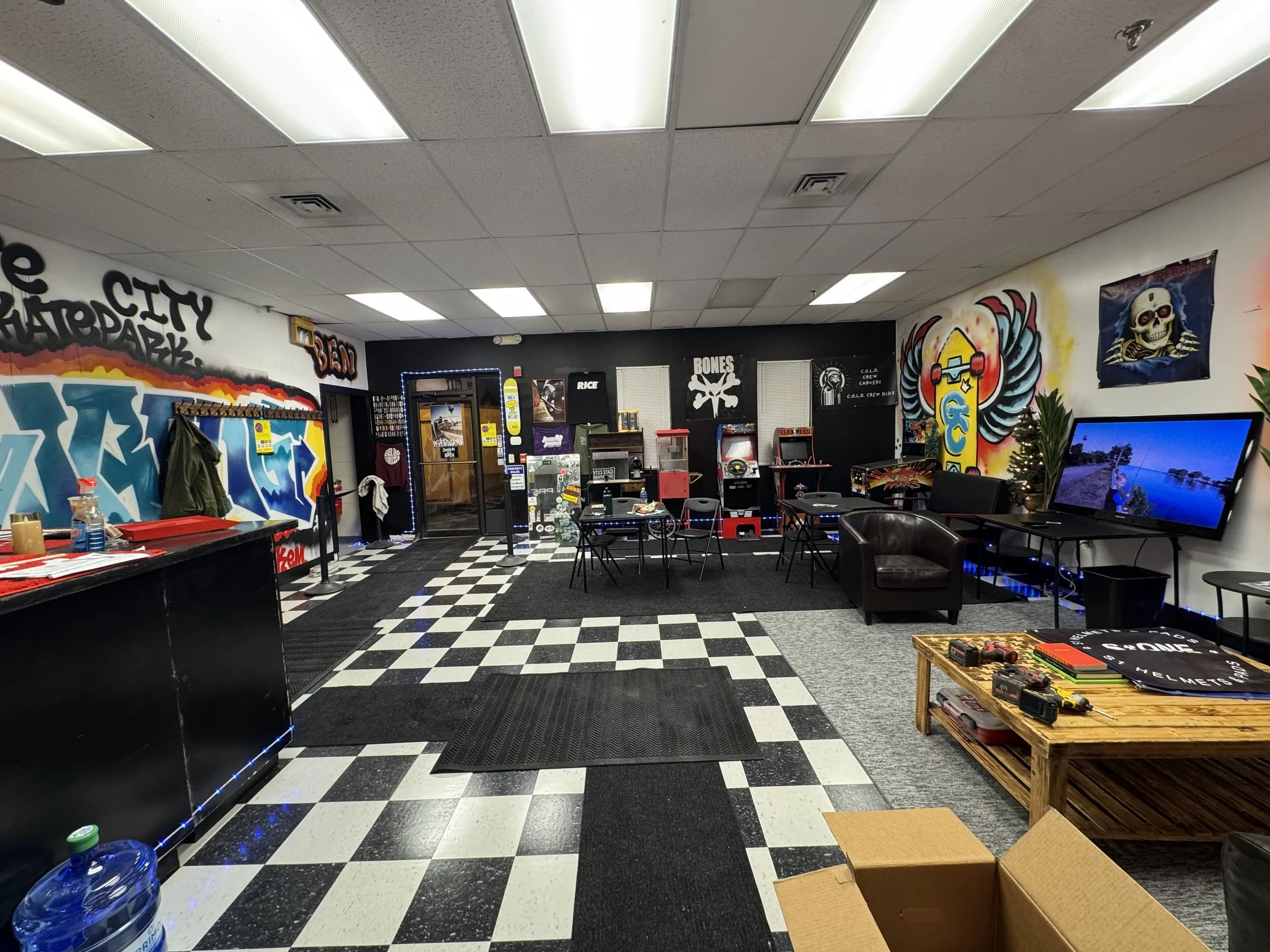 Interior of a game arcade or recreational space with black and white checkered flooring, graffiti-style wall art, arcade machines, a TV screen, a brown couch, a wooden table with toys or tools, and various gaming and themed decorations.